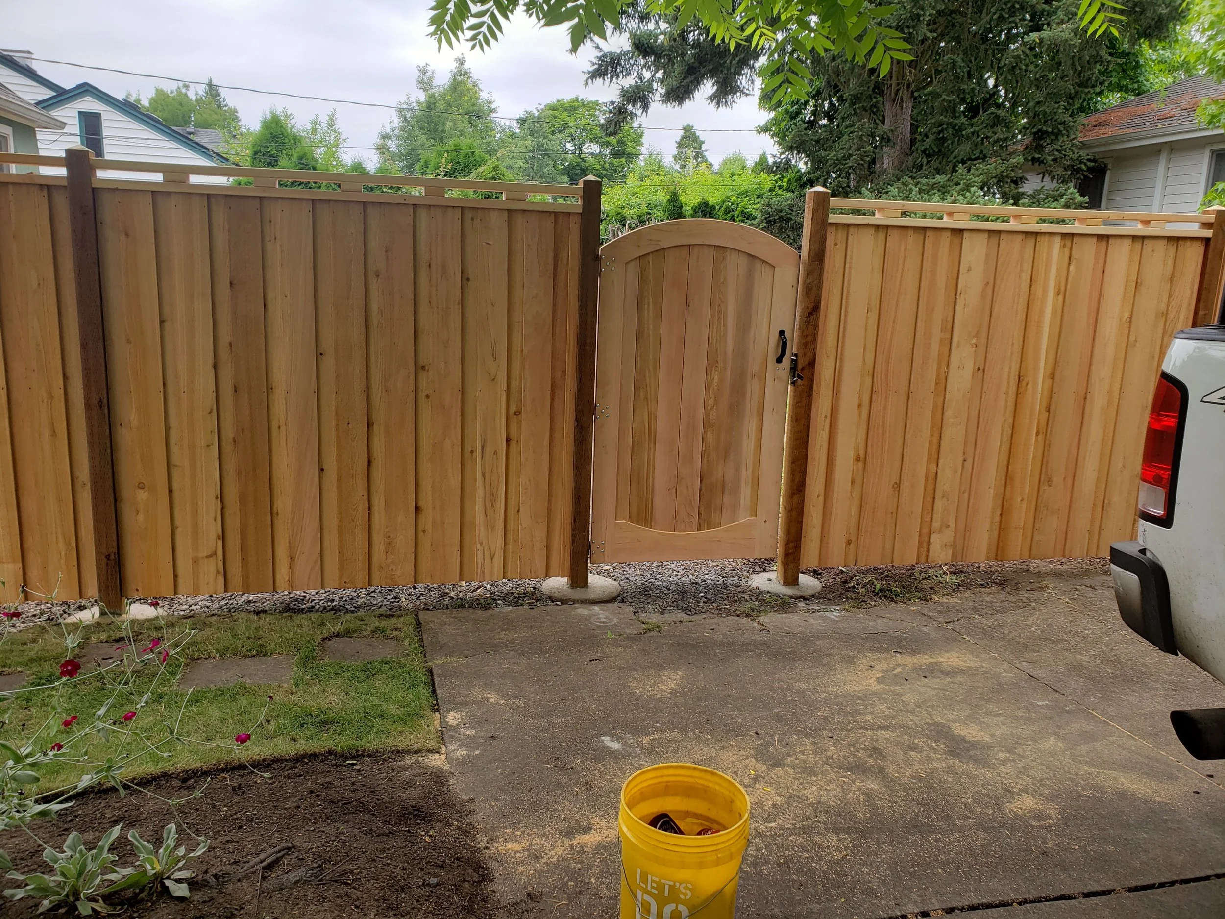 New wooden fence with a small gate, concrete driveway, parked white truck, yellow bucket, garden with plants and flowers, trees, and neighboring houses in the background.