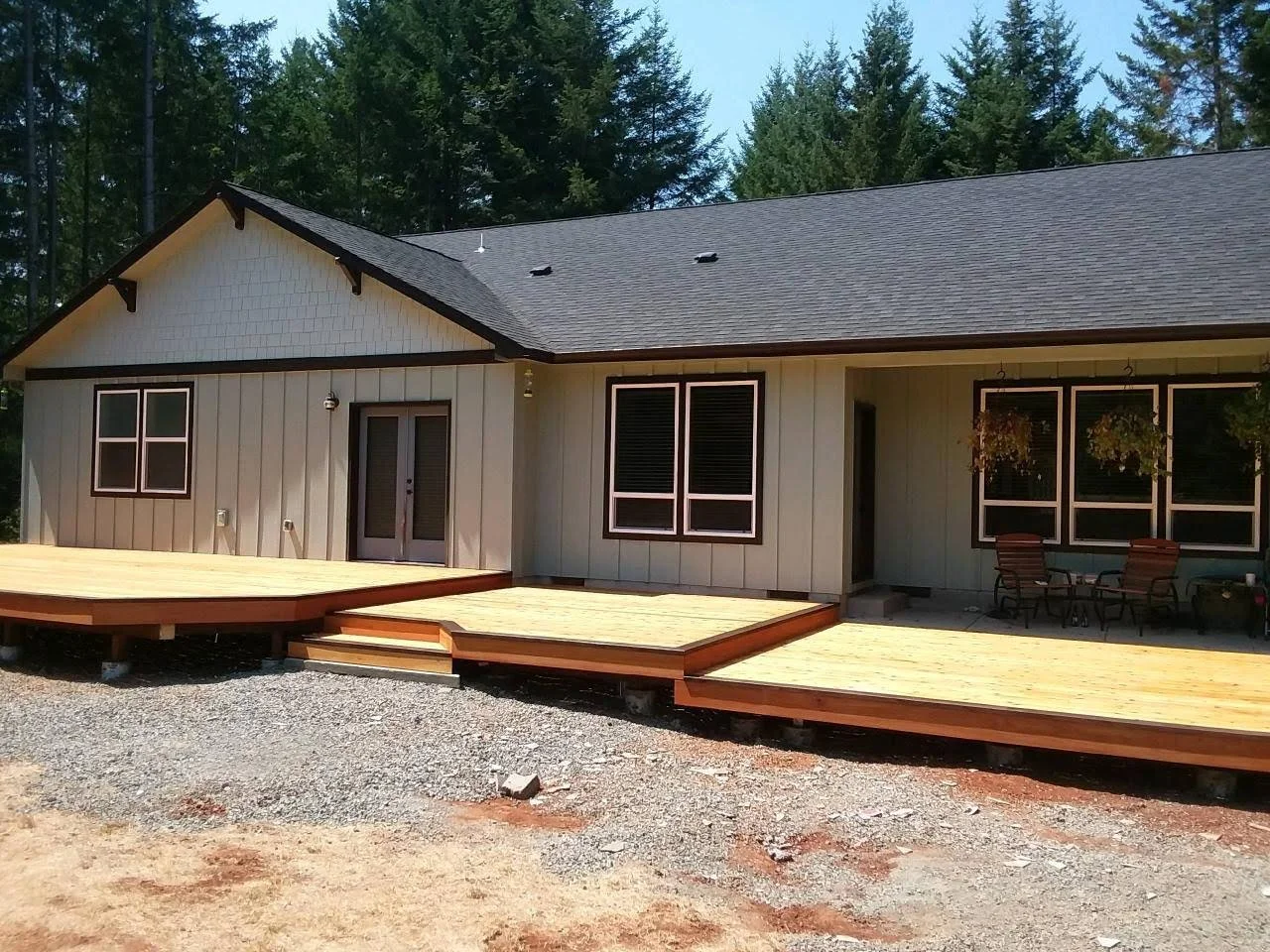 Newly constructed house with a gray roof and beige siding, featuring a wooden deck with steps, surrounded by trees.