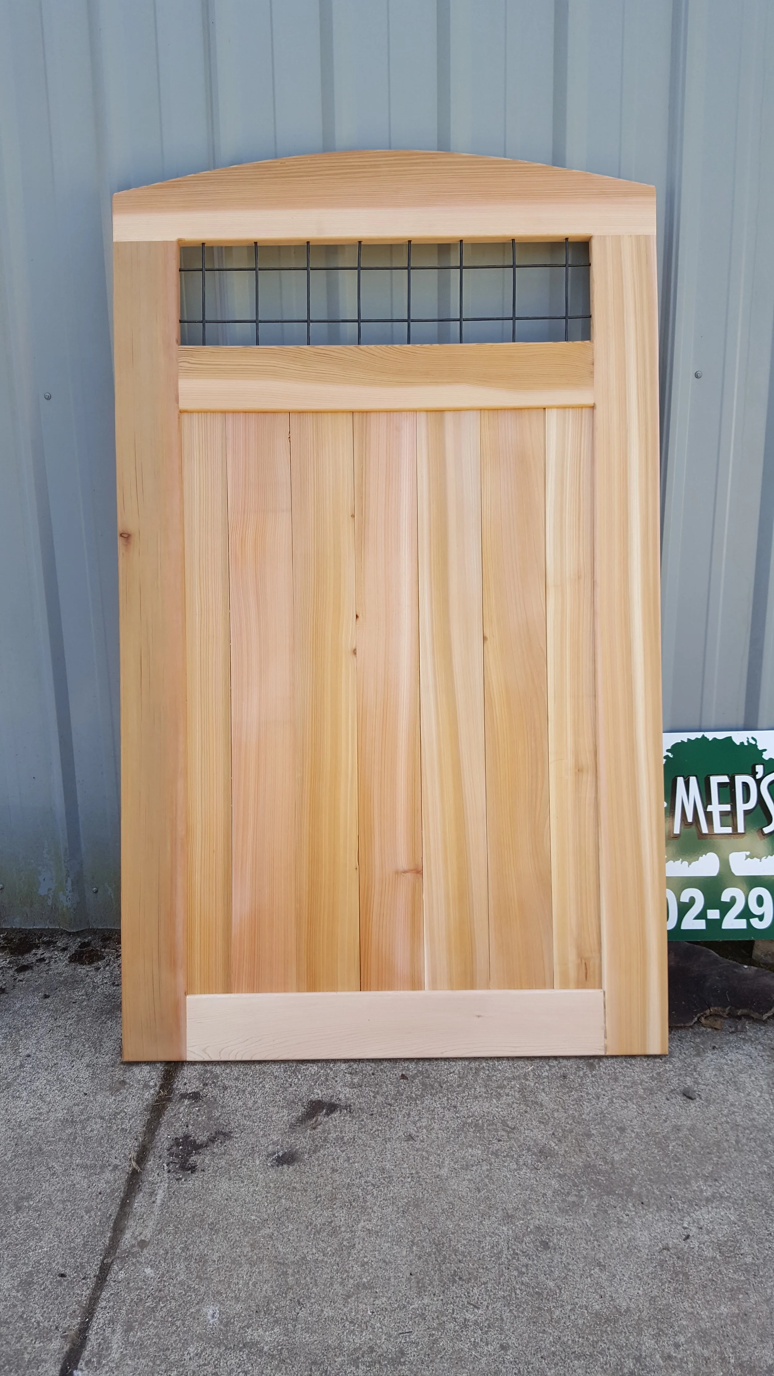 A wooden door with a wire grid at the top, leaning against a metal wall on a concrete surface.