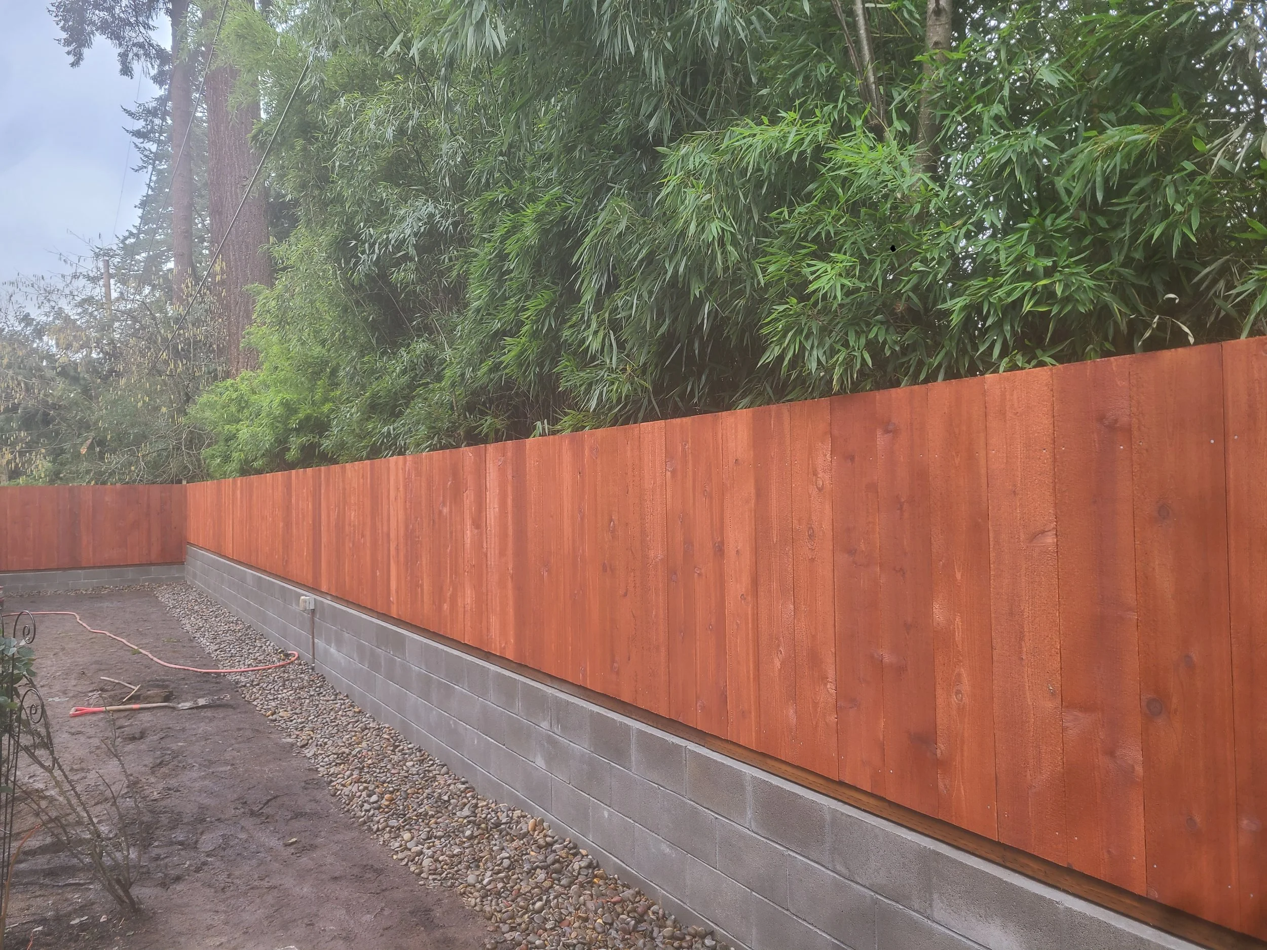 A newly installed wooden fence with vertical reddish-brown planks along a concrete block wall, with trees and greenery in the background.