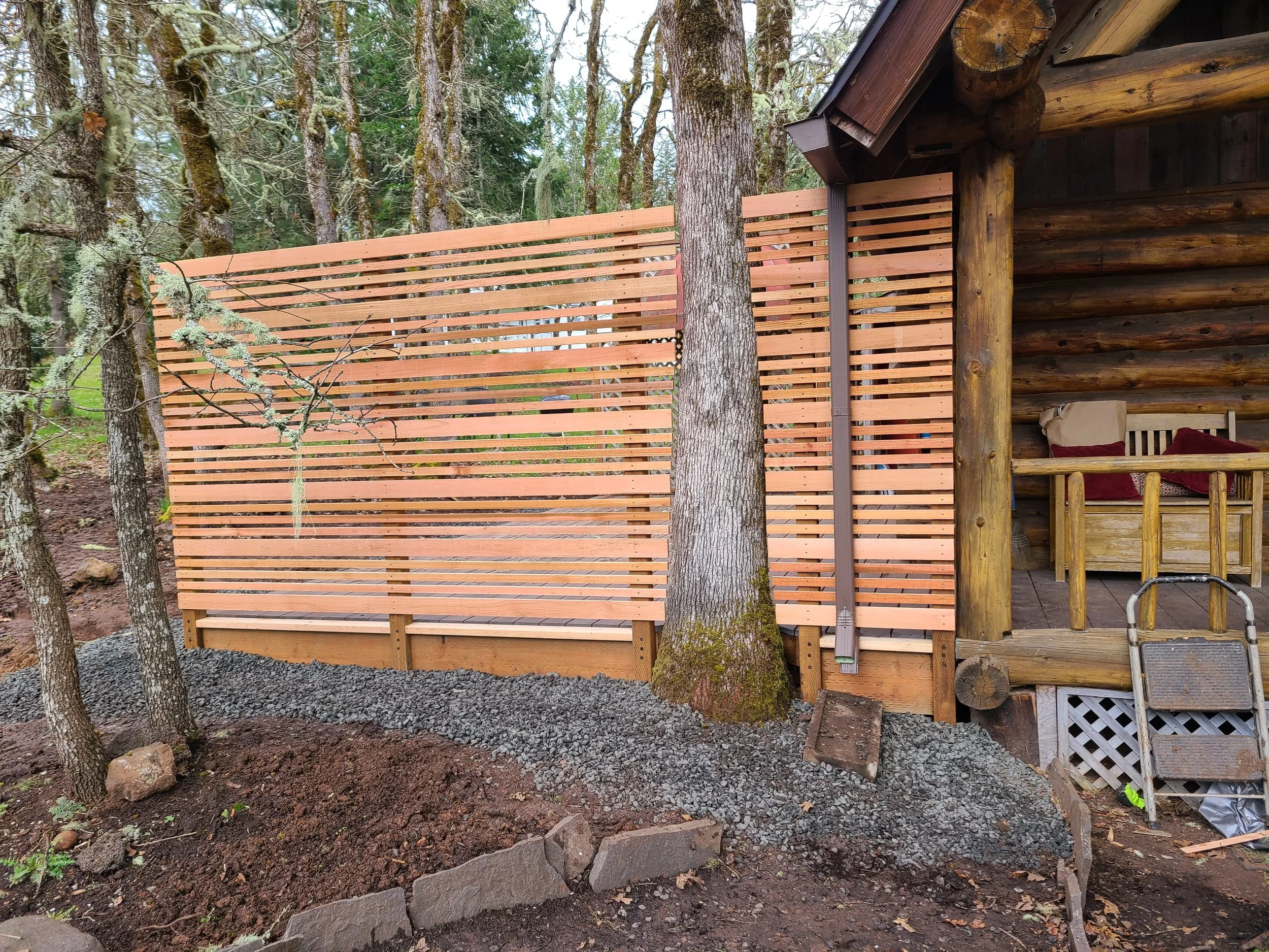 Wooden privacy screen being installed next to a rustic log cabin in a forested area.