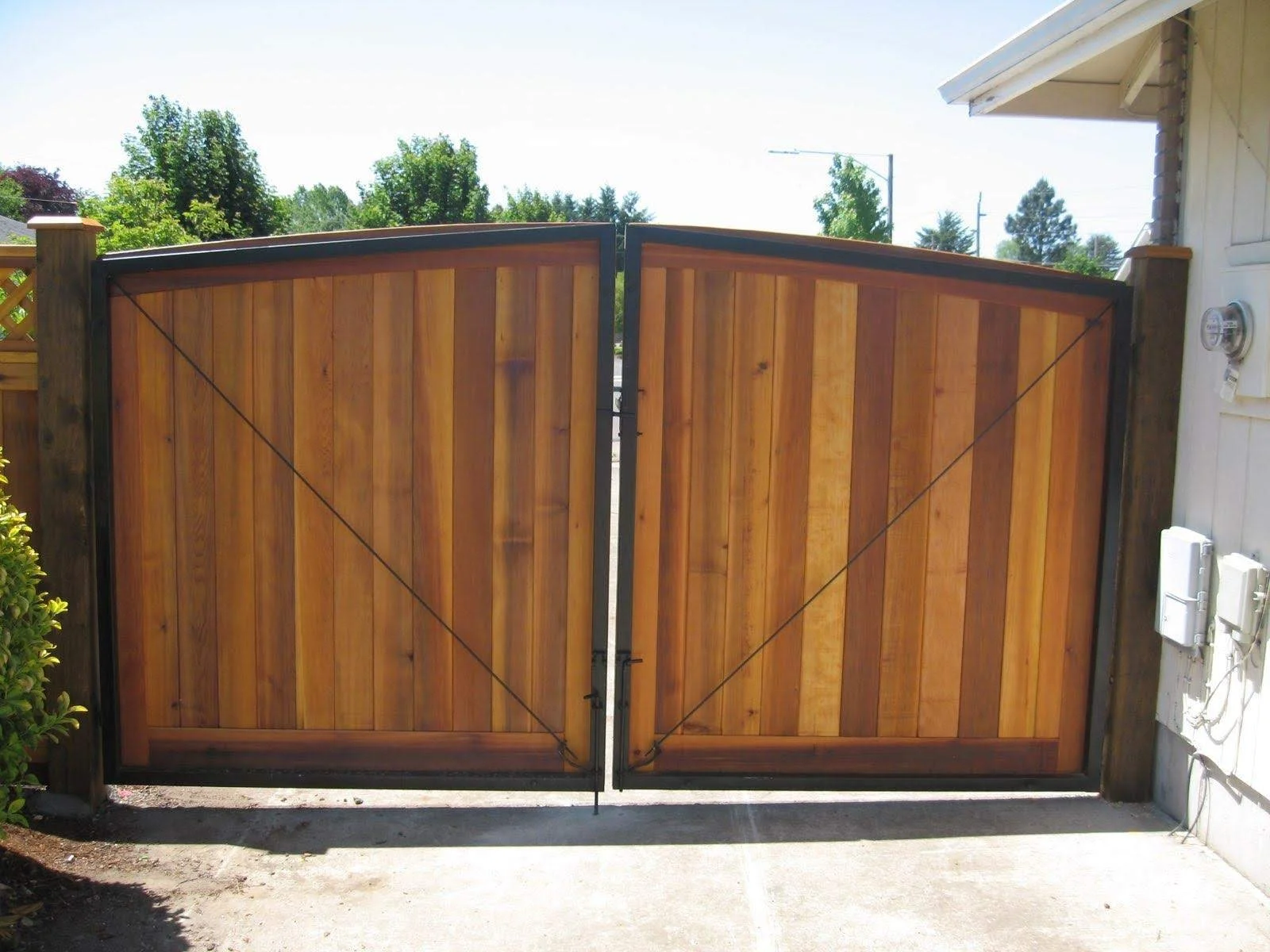 Double wooden gate with diagonal support wires, attached to white house with electrical meters on exterior wall, on a concrete driveway with garden shrub on left and trees in background.