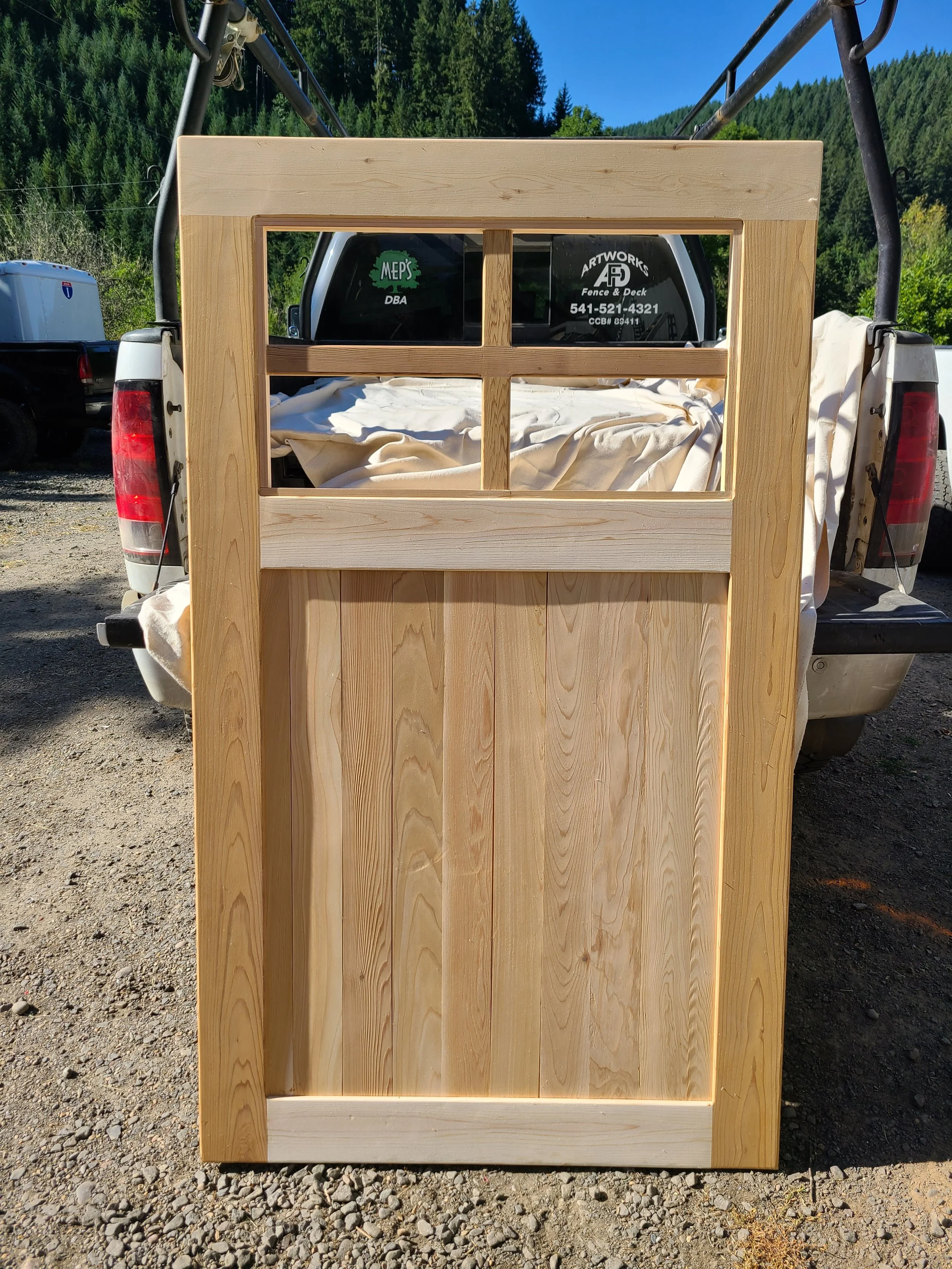 Unfinished wooden door with three window panes at the top, standing upright outside on gravel with a pickup truck and green forest in the background.