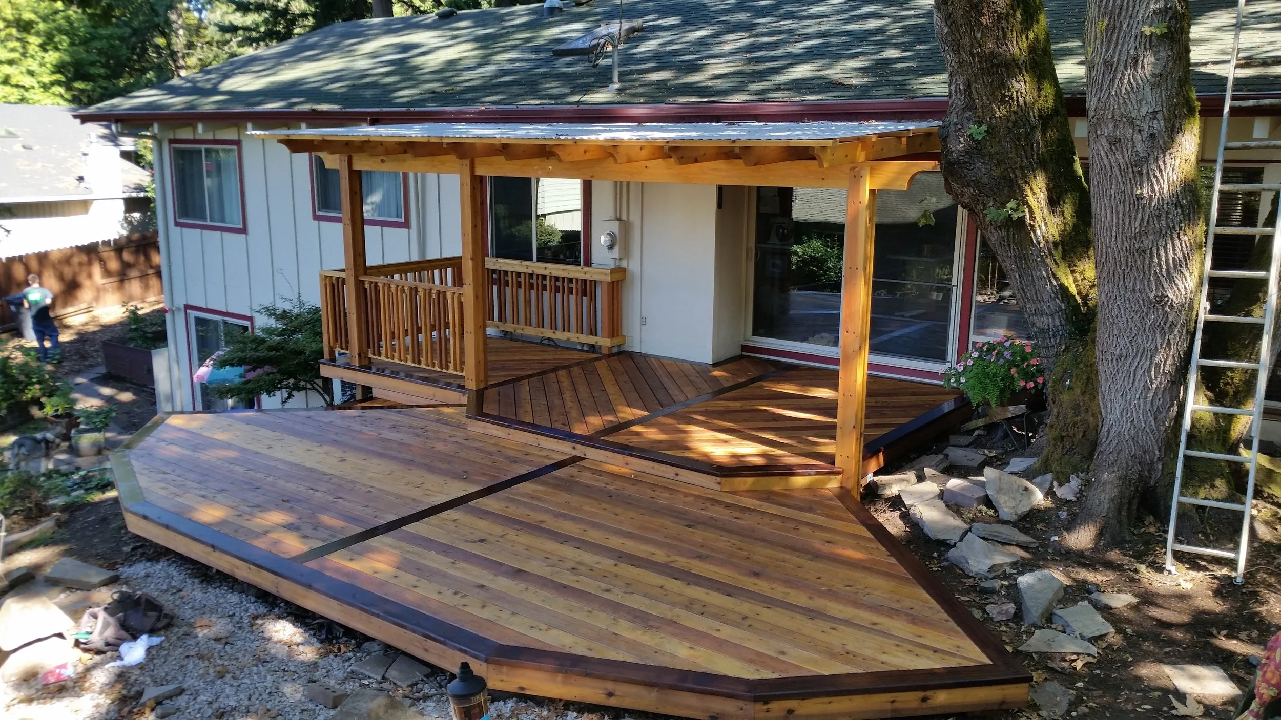 The image shows a newly constructed wooden deck attached to a house, with a partially shaded roof extension. The deck has multiple levels, with the main level featuring a wooden railing and smaller platforms. The house has a white exterior with large sliding glass doors and several windows. Two mature trees are adjacent to the deck, and a ladder is leaning against one of them. In the background, there is a person working in the yard and some gardening equipment.