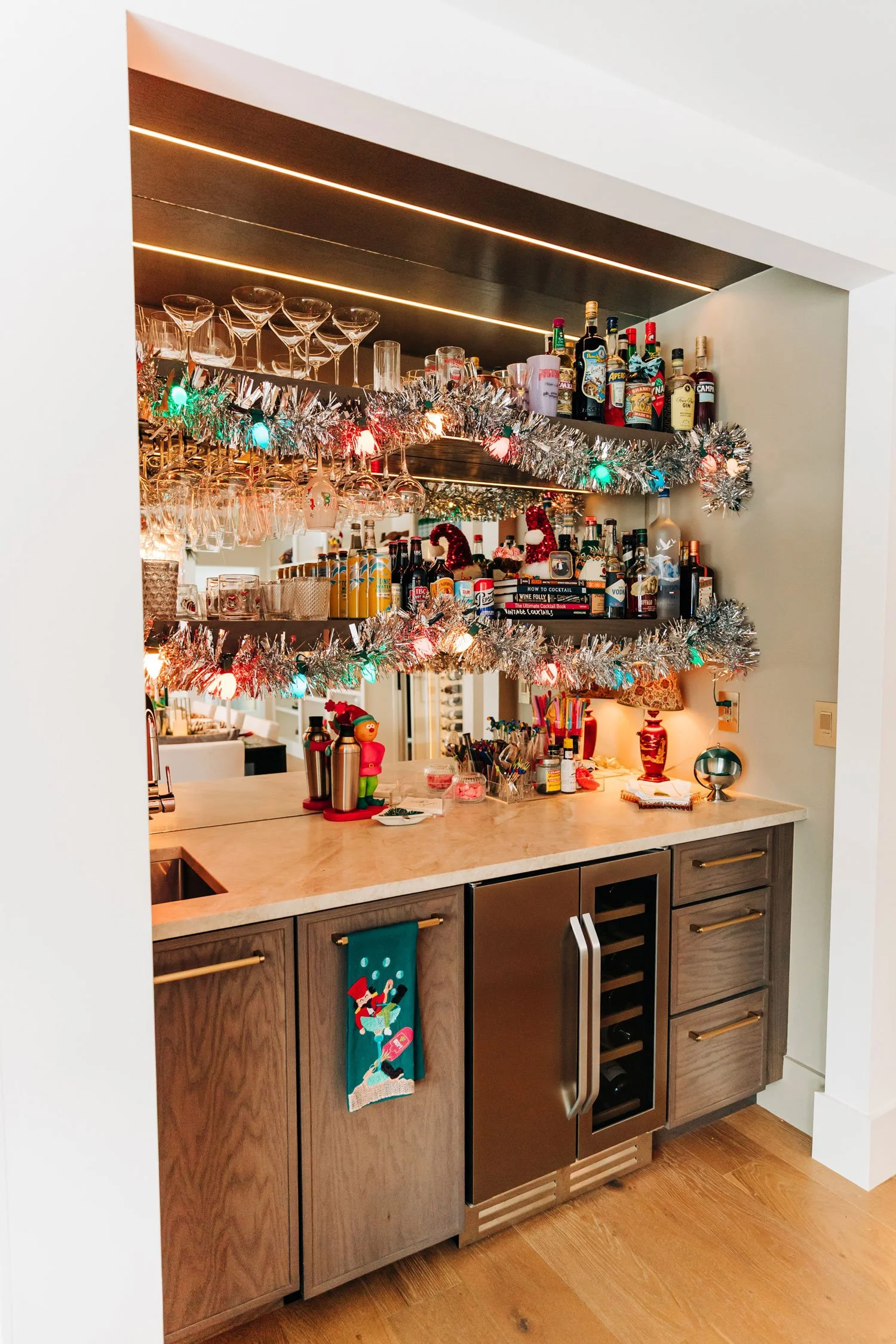 Home bar styled like a party with tinsel garland, hanging glassware, and mixers set out for guests to serve themselves