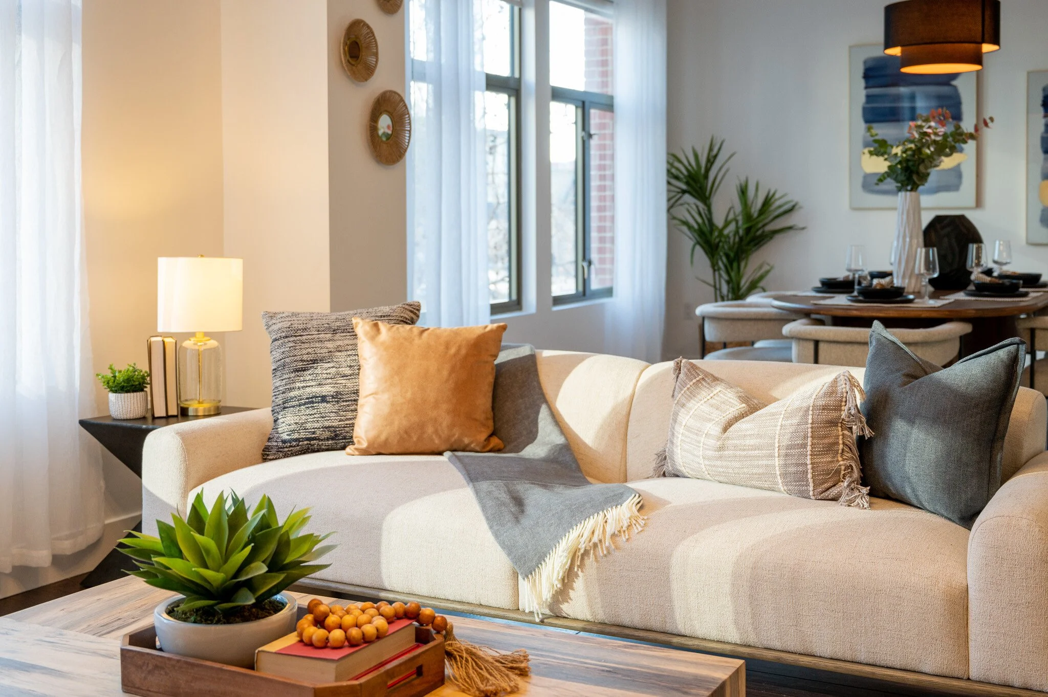 Modern living room with beige sofa, decorative pillows, and a coffee table featuring books and a plant. A dining area with a round table is visible in the background, along with wall art and natural light streaming through windows.