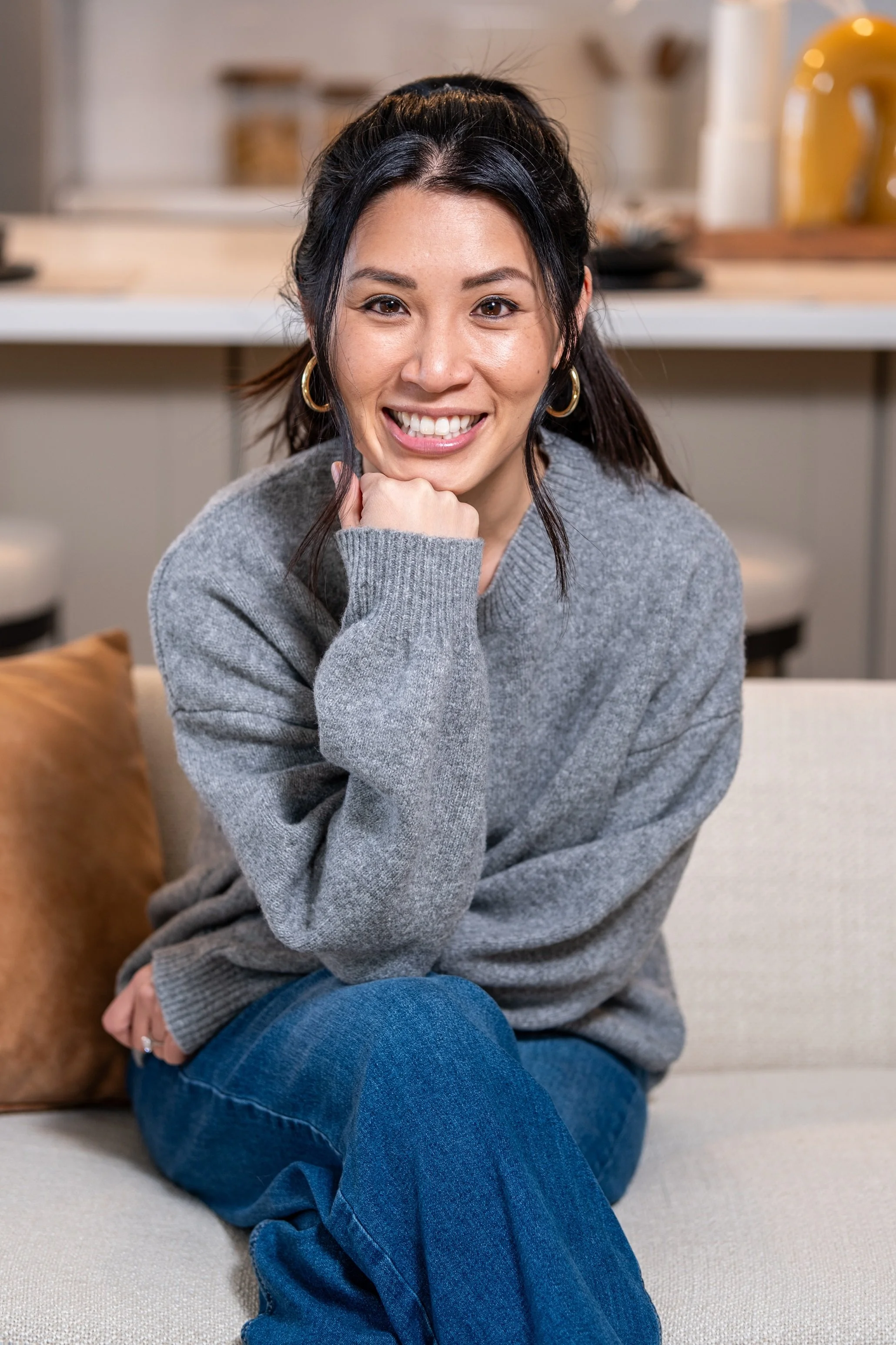 Woman smiling, sitting on a couch, wearing a gray sweater and jeans.
