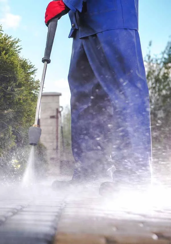 Person pressure washing a wooden deck outdoors, with water spray visible and green bushes on the side.