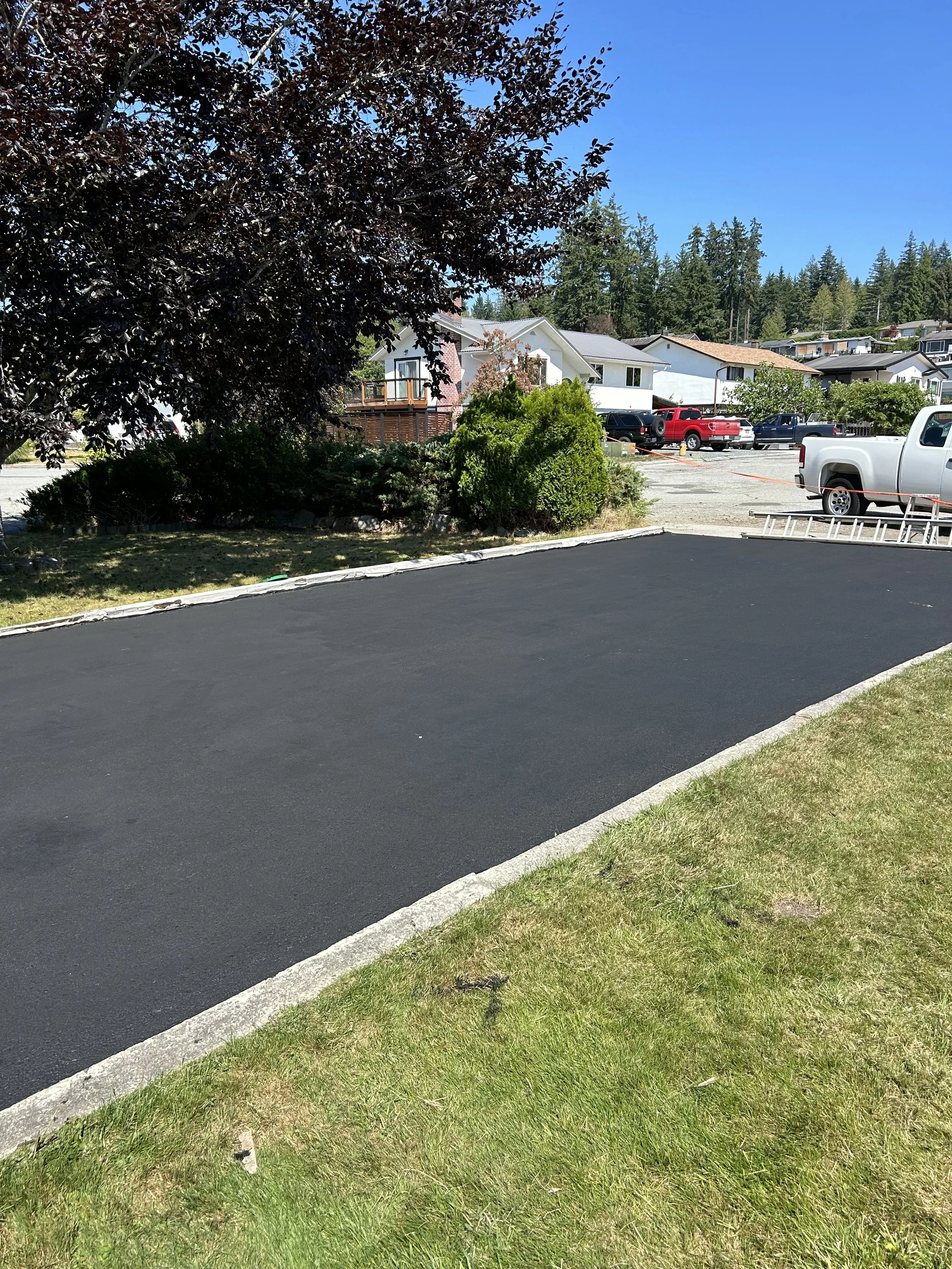 A freshly paved blacktop parking space with a grassy area in the foreground, trees, and a residential neighborhood with parked trucks and houses in the background.