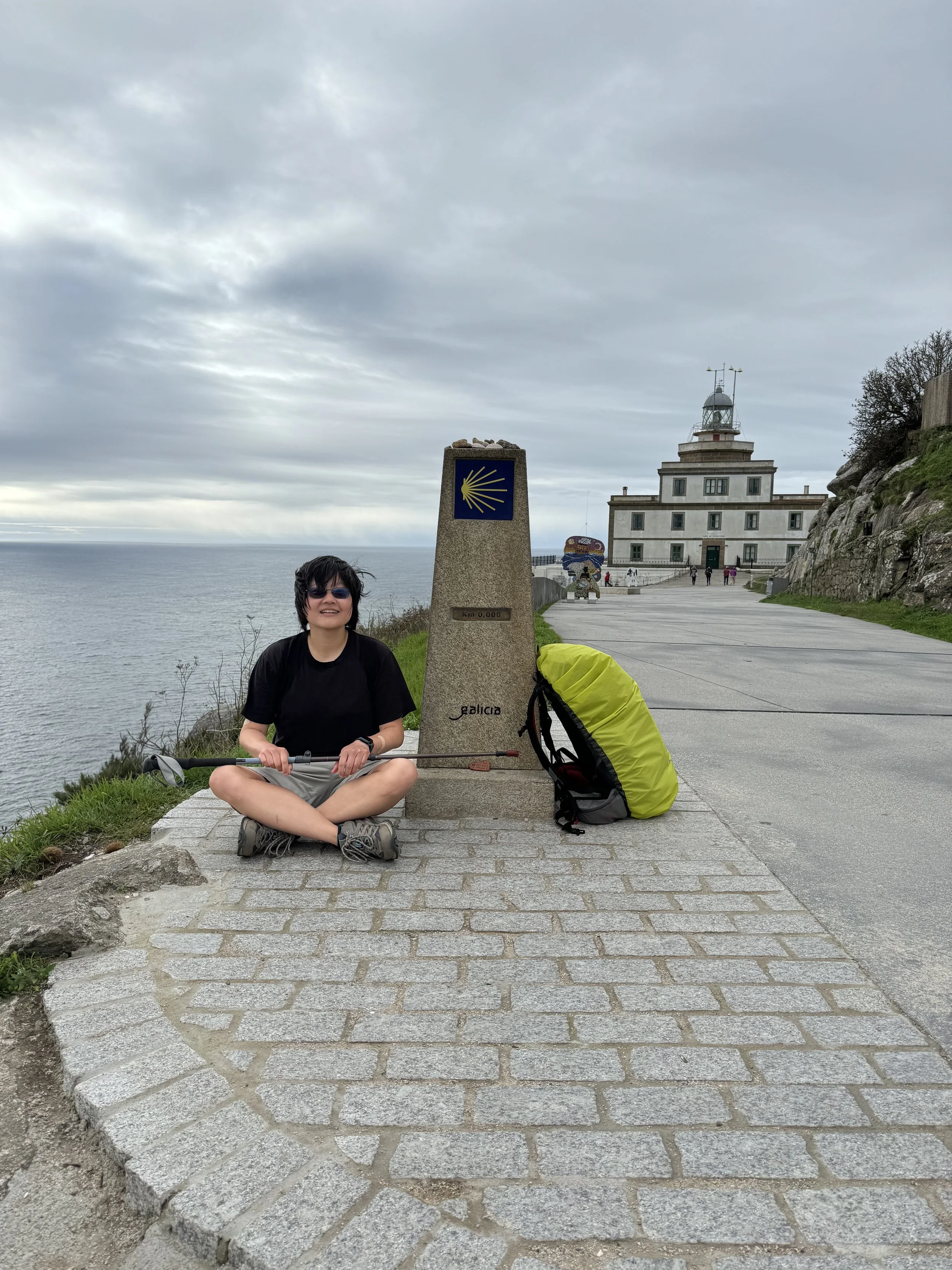 Person sitting cross-legged on a walkway beside a Camino de Santiago marker post, with a yellow shell symbol, near a coast. A yellow and gray backpack lies nearby. A building in the background with a cloudy sky overhead.