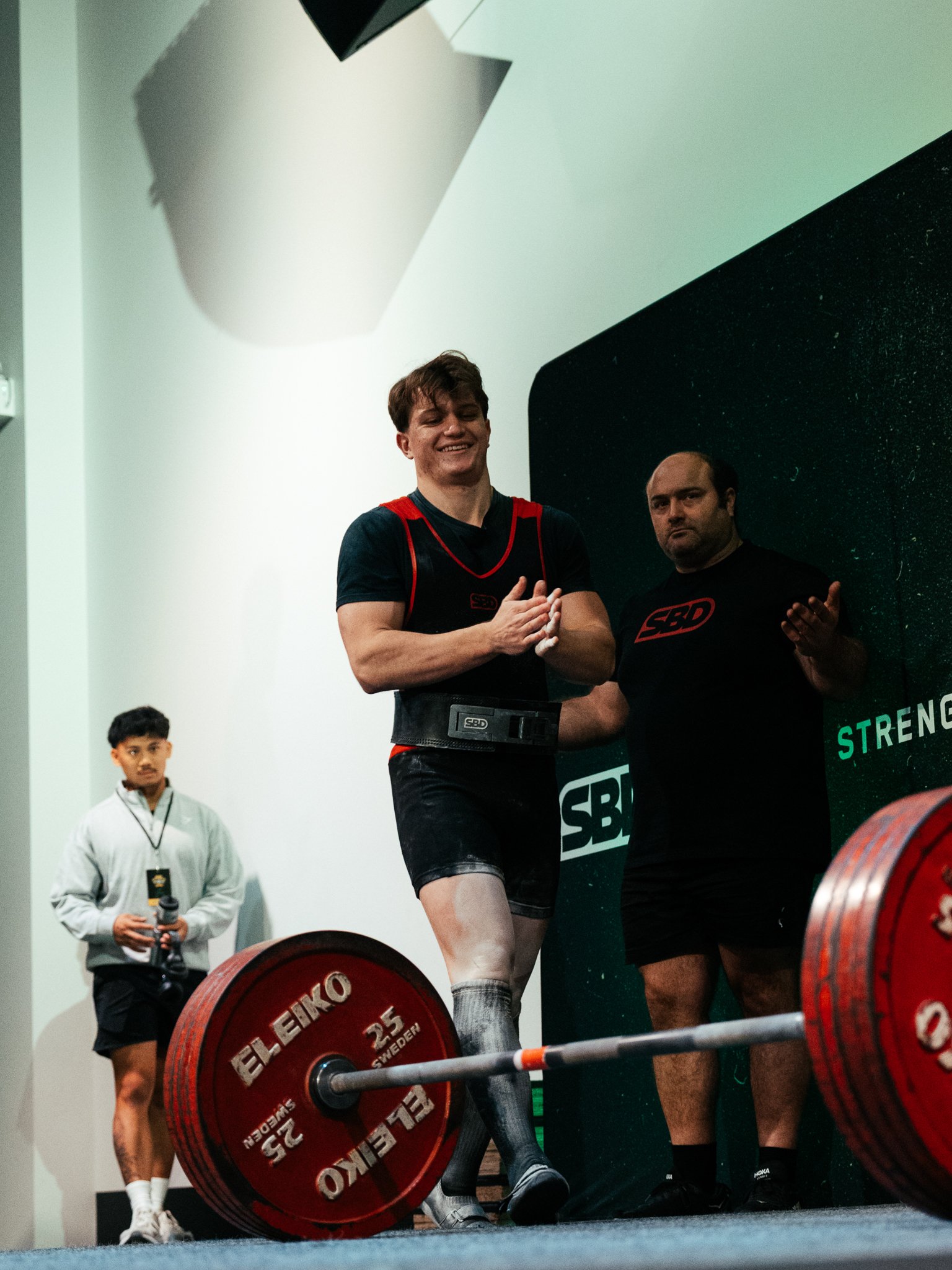 Young man lifting a heavily loaded barbell at a powerlifting competition, with a person in the background and others nearby observing.