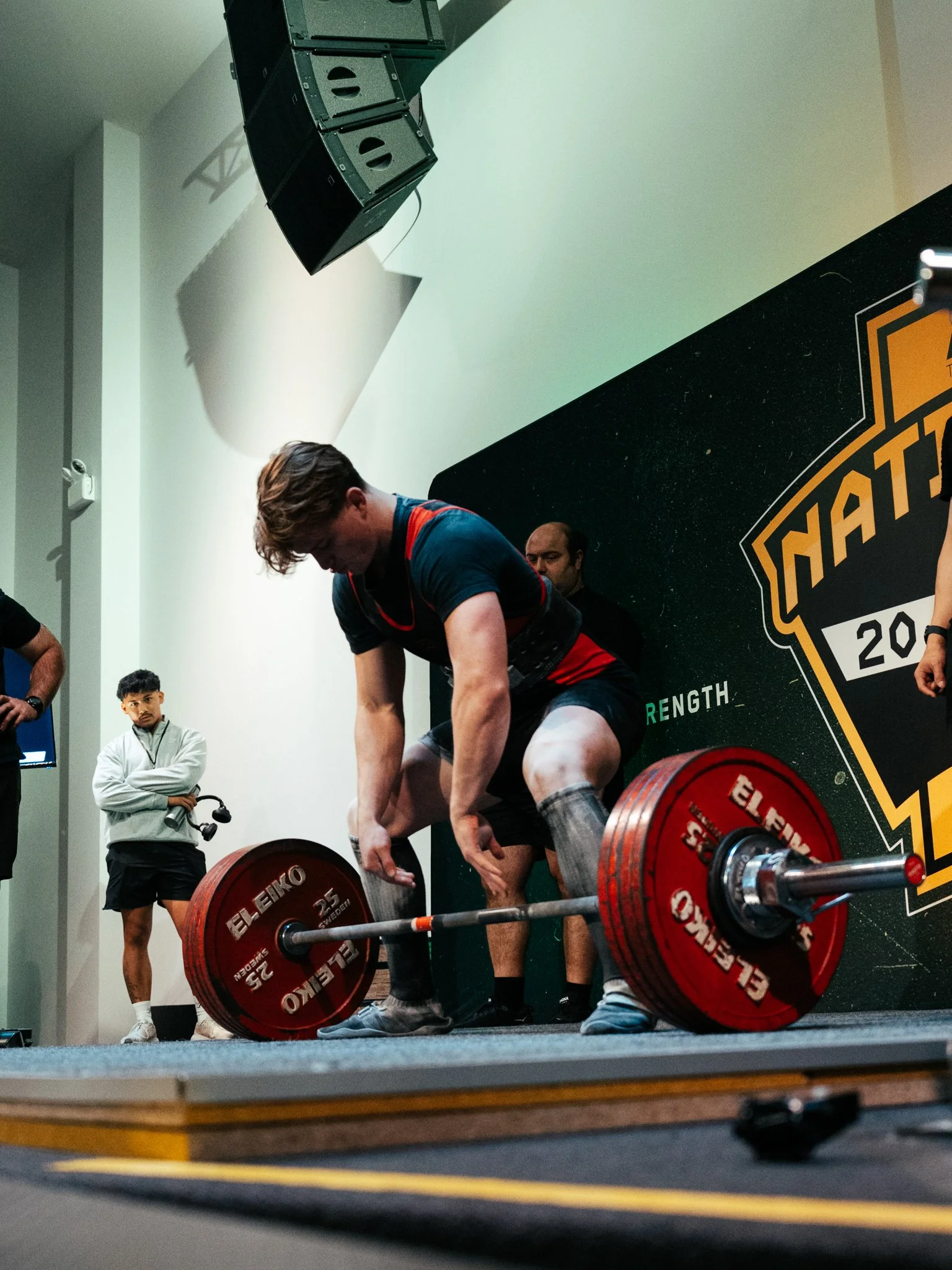 A man is preparing to lift a loaded barbell during a powerlifting competition, with a coach or judge standing nearby, observing, and a young spectator watching from the background.