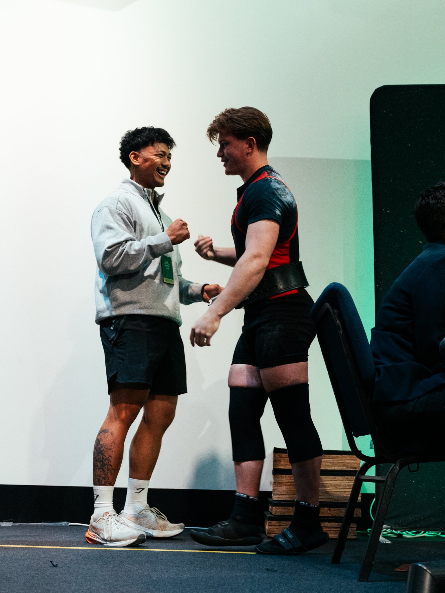 Two men talking and smiling, standing on a stage, one wearing athletic clothes and the other in black shorts, with chairs and equipment nearby.