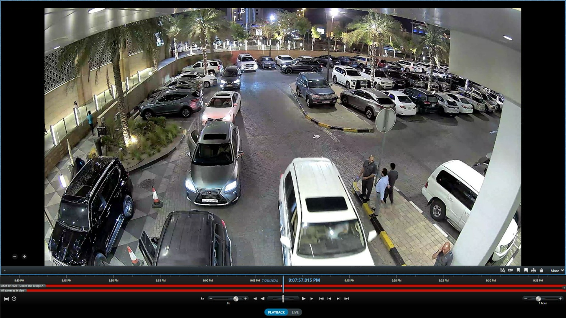 Nighttime view of a parking lot filled with various parked cars and a few pedestrians walking along the sidewalk.