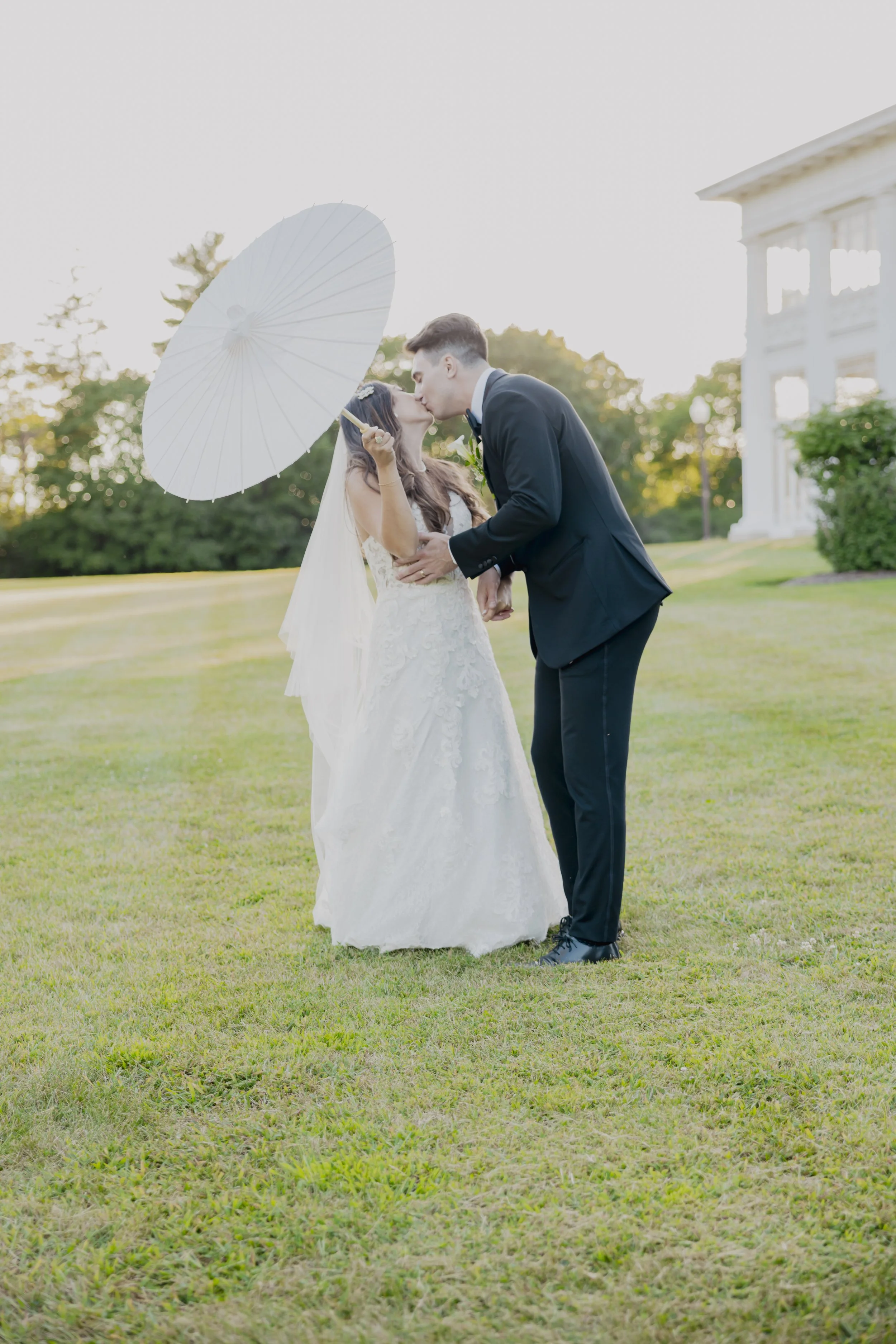 A bride and groom kissing outside on a grassy lawn during their wedding, with the bride holding a white parasol.