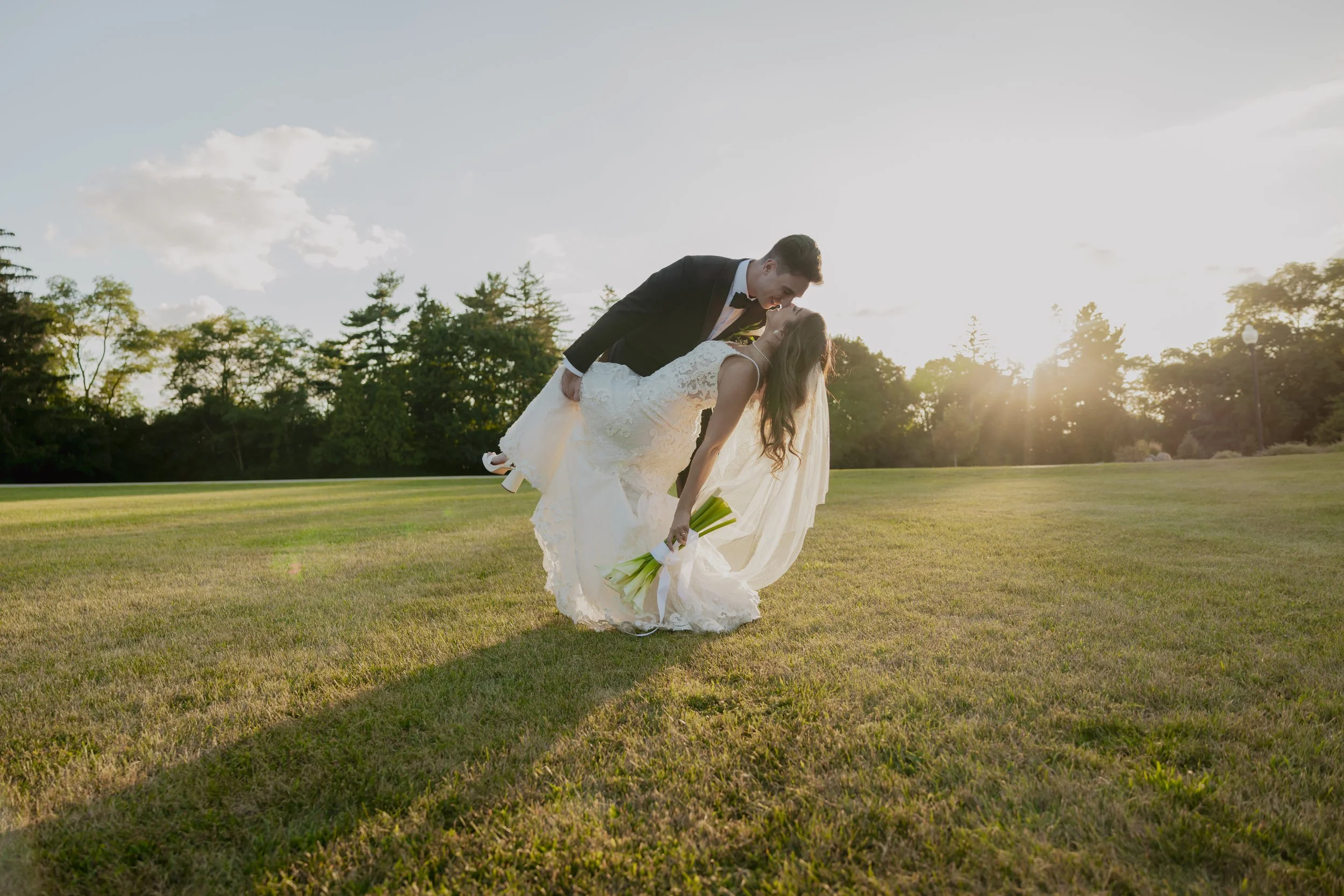 A bride and groom kissing in a field during sunset, with the groom dipping the bride and she holding a bouquet.