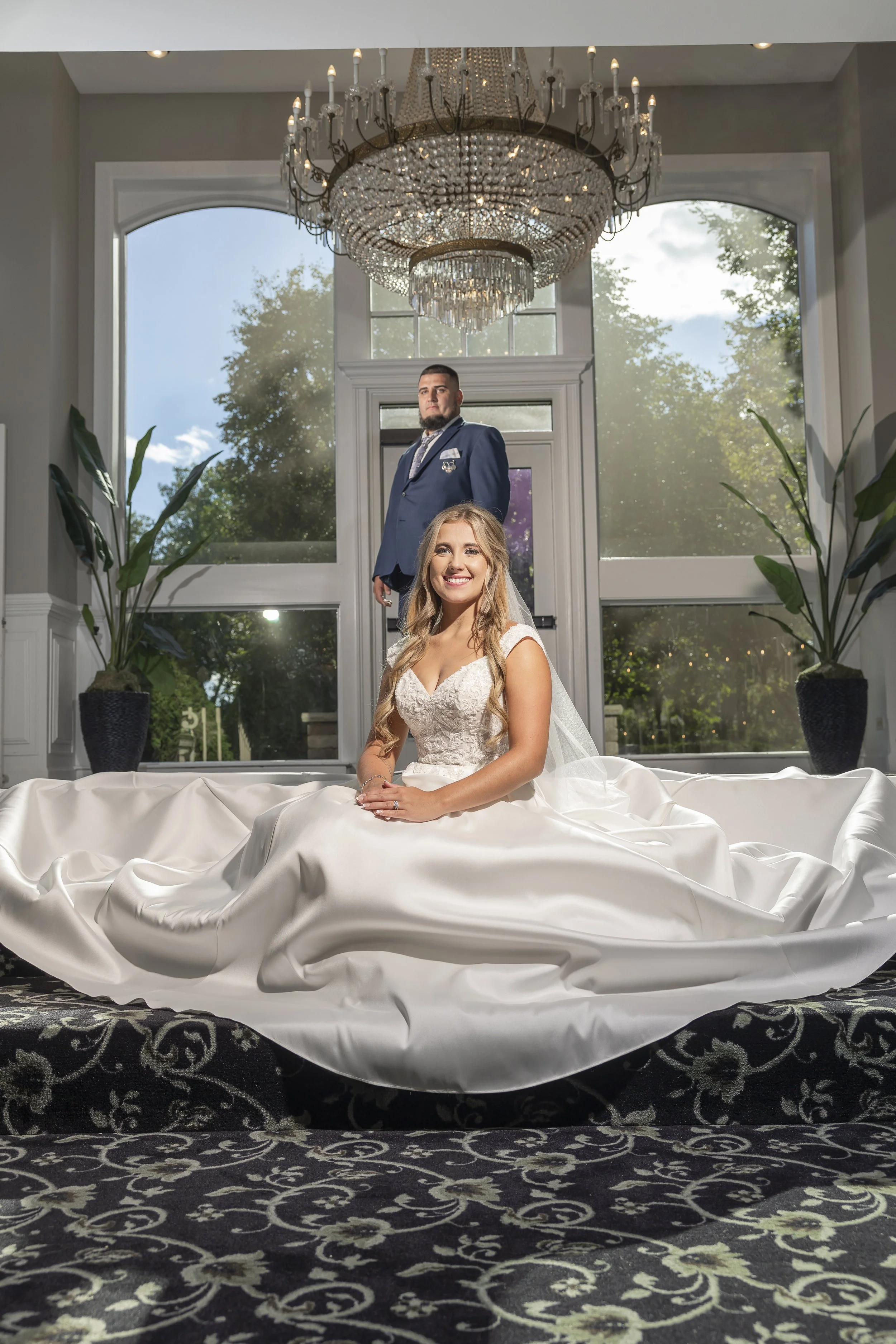 Bride in a white wedding gown sitting on a luxurious black and white patterned rug, smiling, with a groom in a blue suit standing behind her in front of large windows and a chandelier.