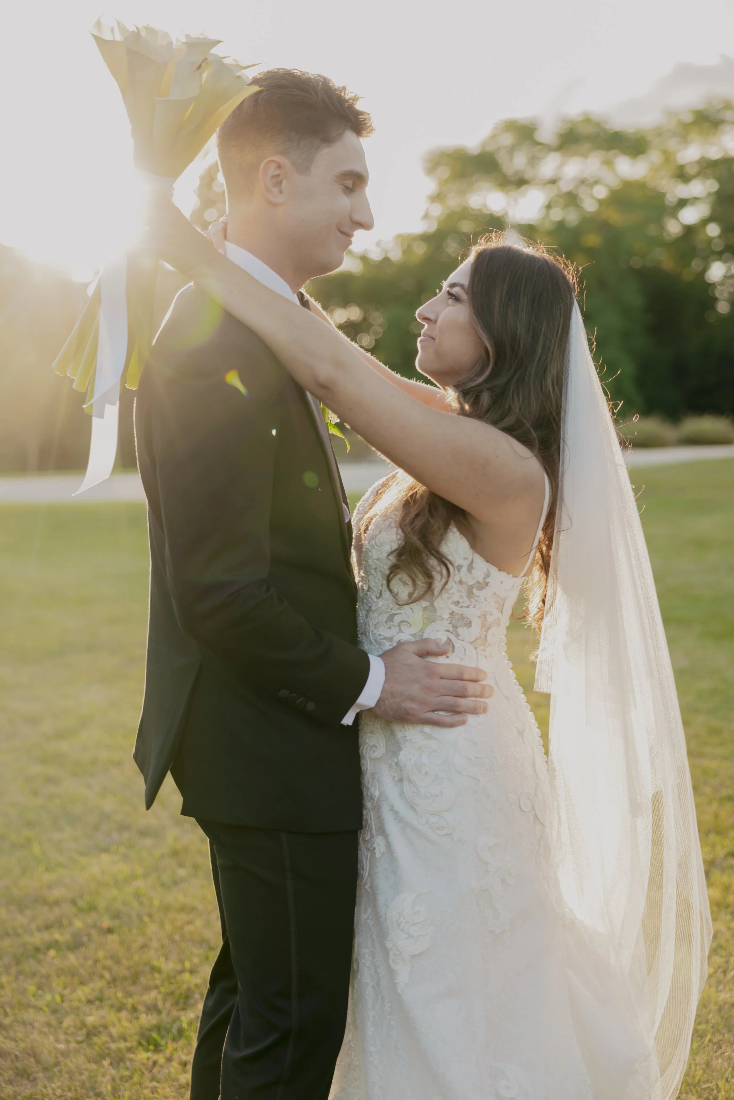 A bride and groom embracing outdoors during sunset, with the bride holding a bouquet of flowers over the groom's shoulder.