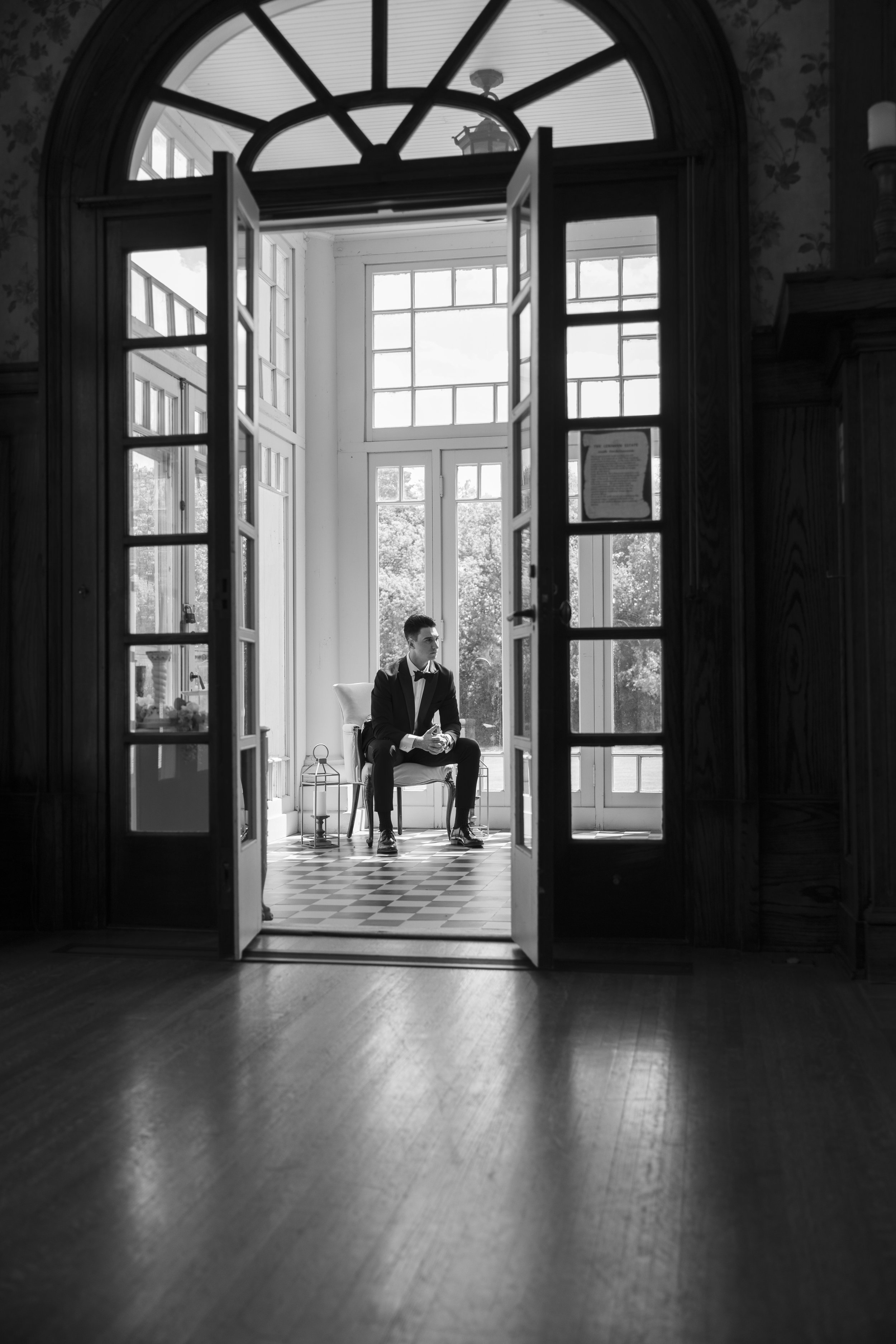 A man in a tuxedo sitting on a chair in a sunlit conservatory with large windows and checkered flooring, viewed through a doorway with open double doors.