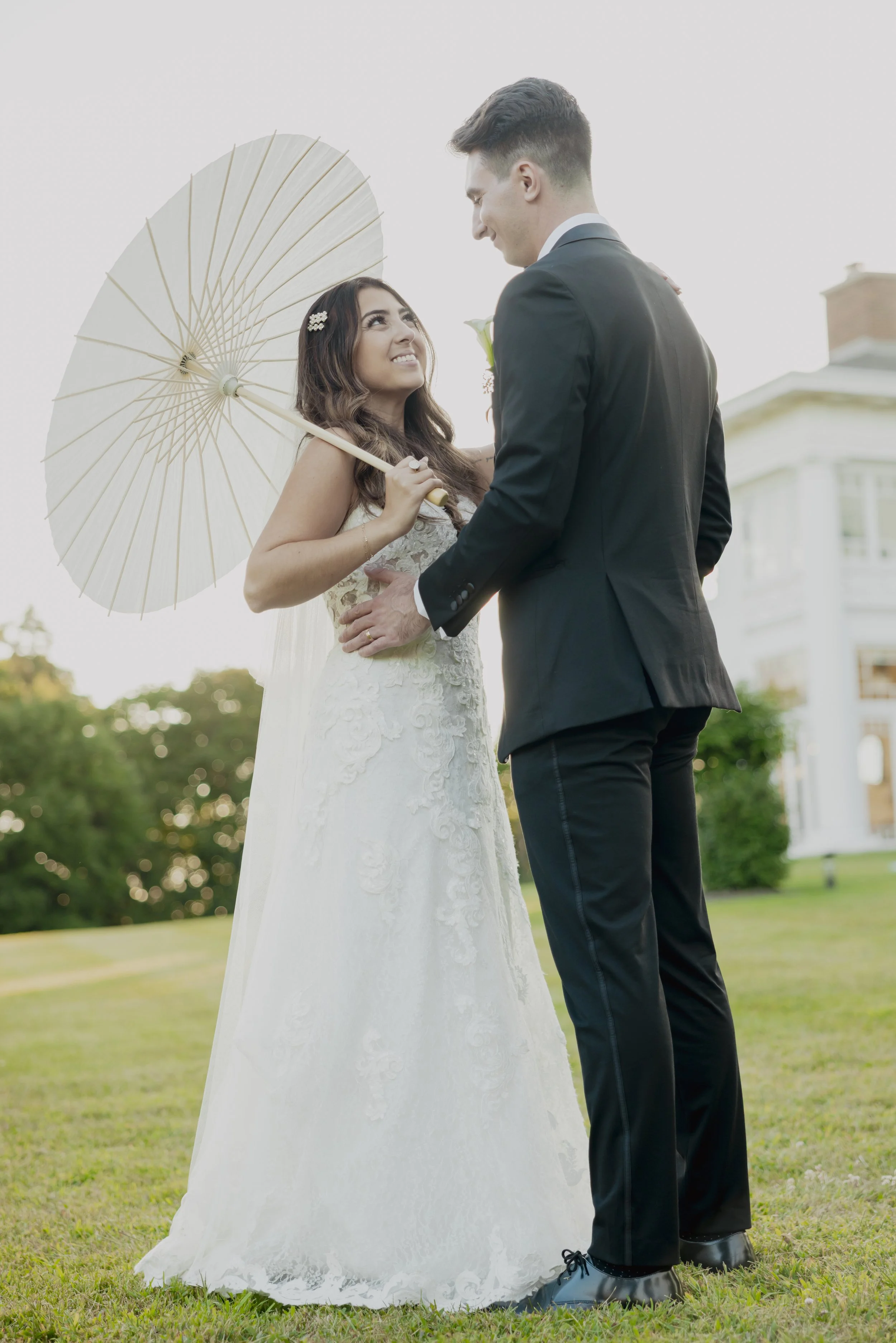 A bride holding a parasol and a groom standing on a lawn, facing each other and smiling, with a large white house in the background.