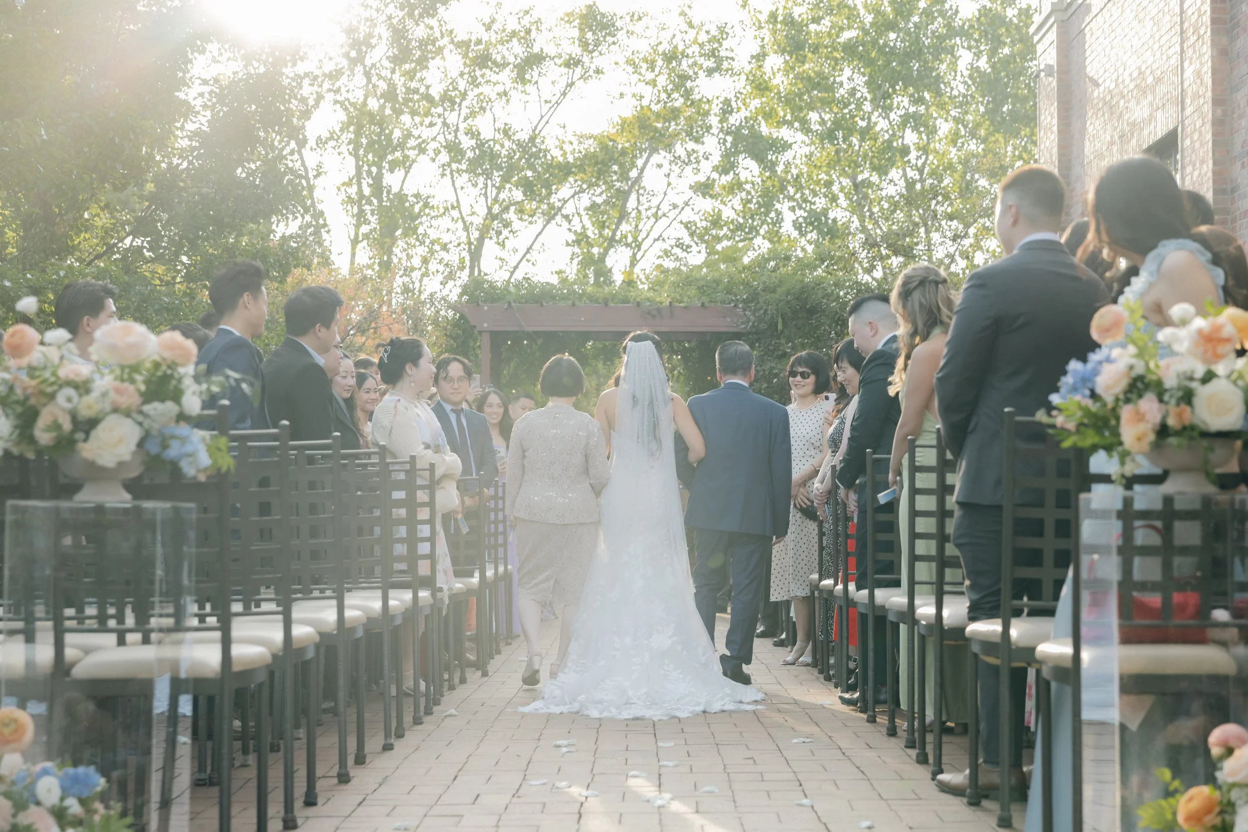 Bride walking down the aisle at an outdoor wedding ceremony, flanked by guests on both sides, with sunlight filtering through trees in the background.