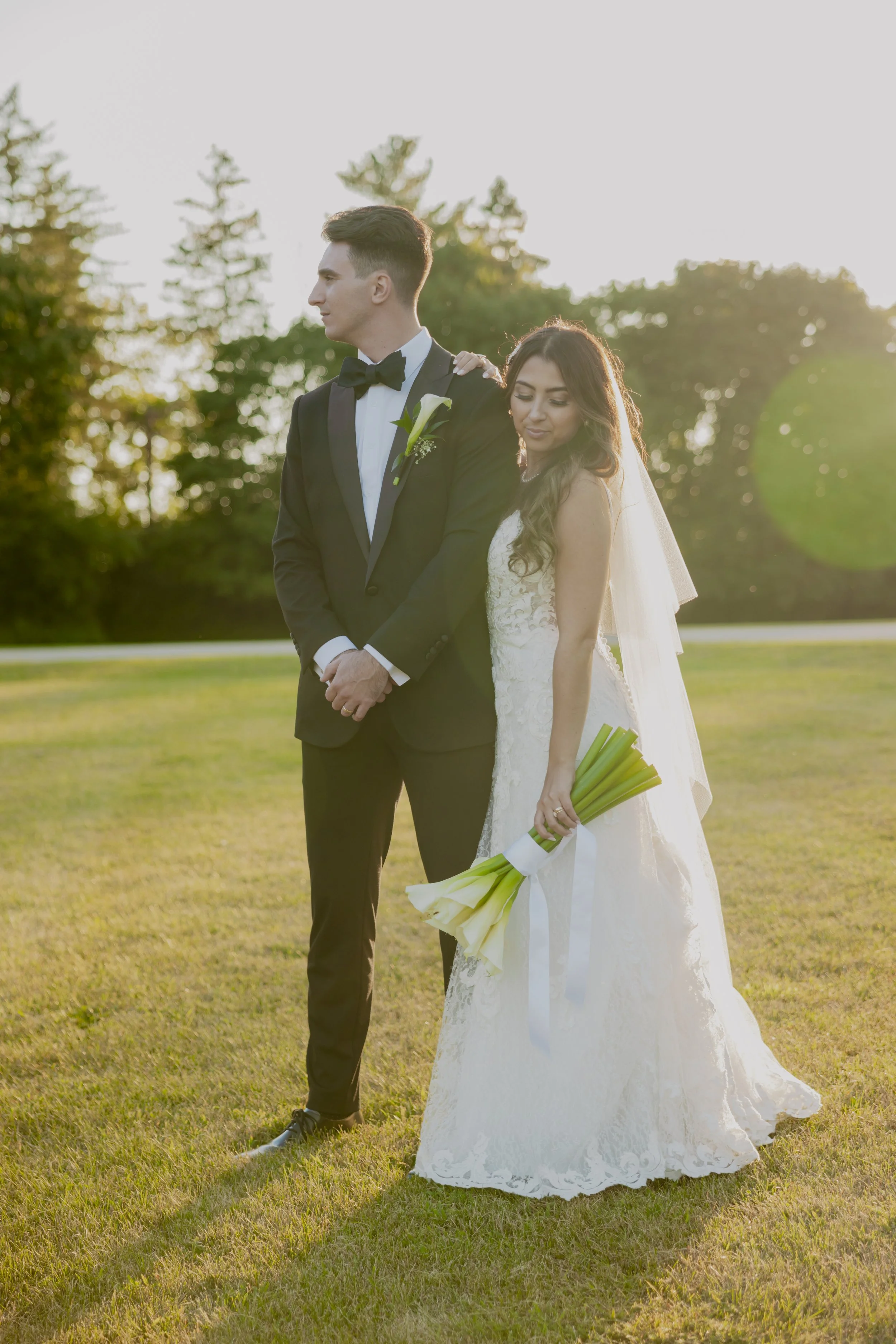 A bride and groom standing on a grassy field during sunset, with the bride holding a bouquet of white calla lilies. The groom wears a black tuxedo with a bow tie, and the bride wears a white lace wedding gown with a veil.