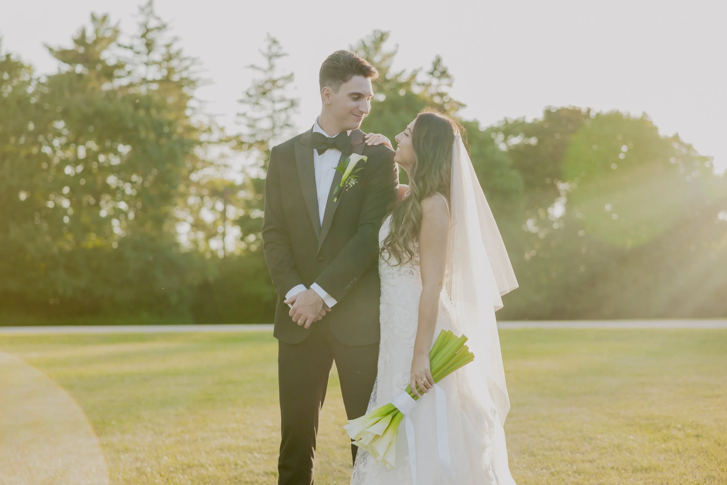 A bride and groom standing outdoors on their wedding day, gazing at each other. The groom is dressed in a black tuxedo with a bow tie and a white boutonniere, while the bride wears a white lace wedding dress and veil, holding a bouquet of lilies. The