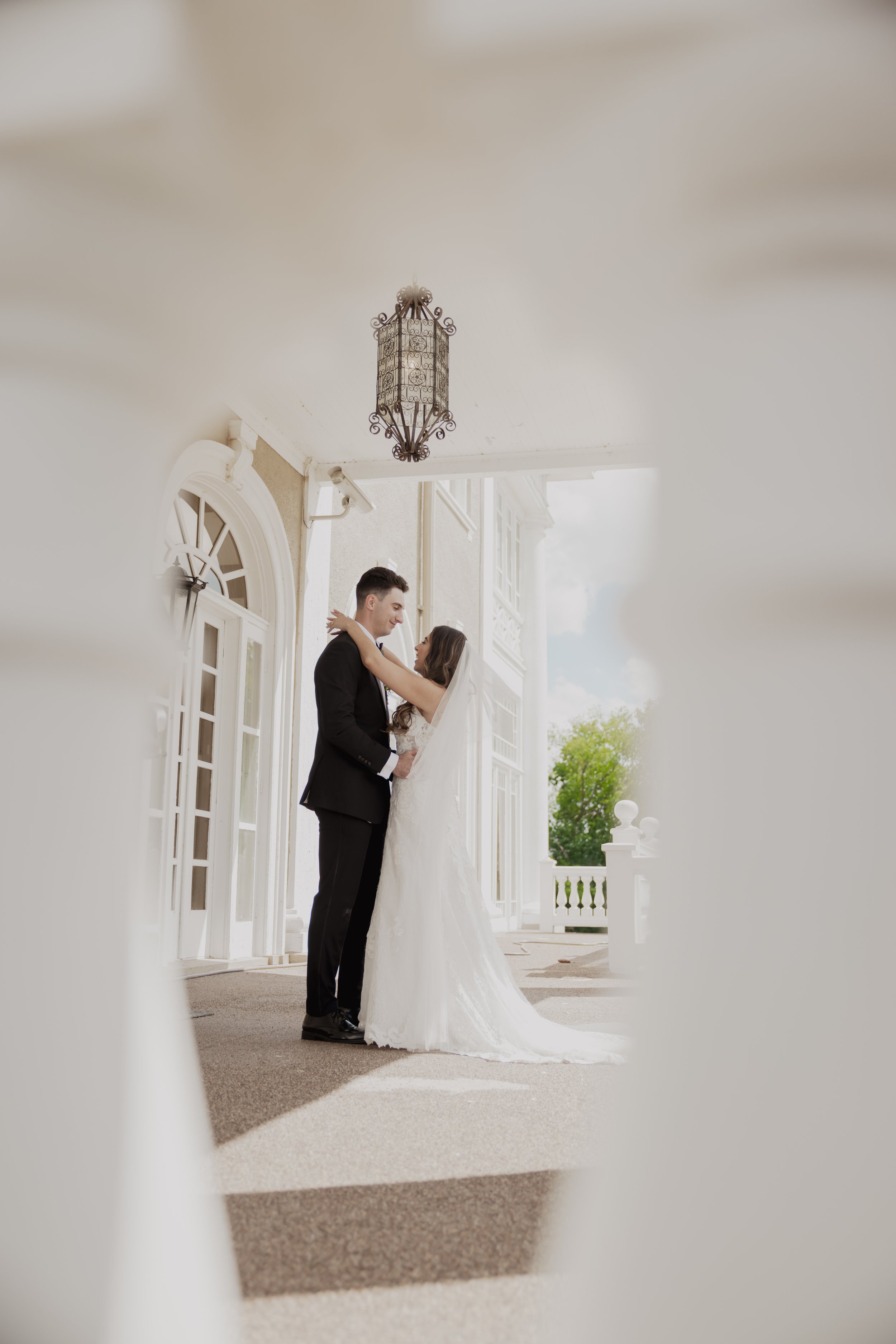 A bride and groom standing close together on a porch, gazing into each other's eyes, surrounded by white architectural elements and a hanging lantern.