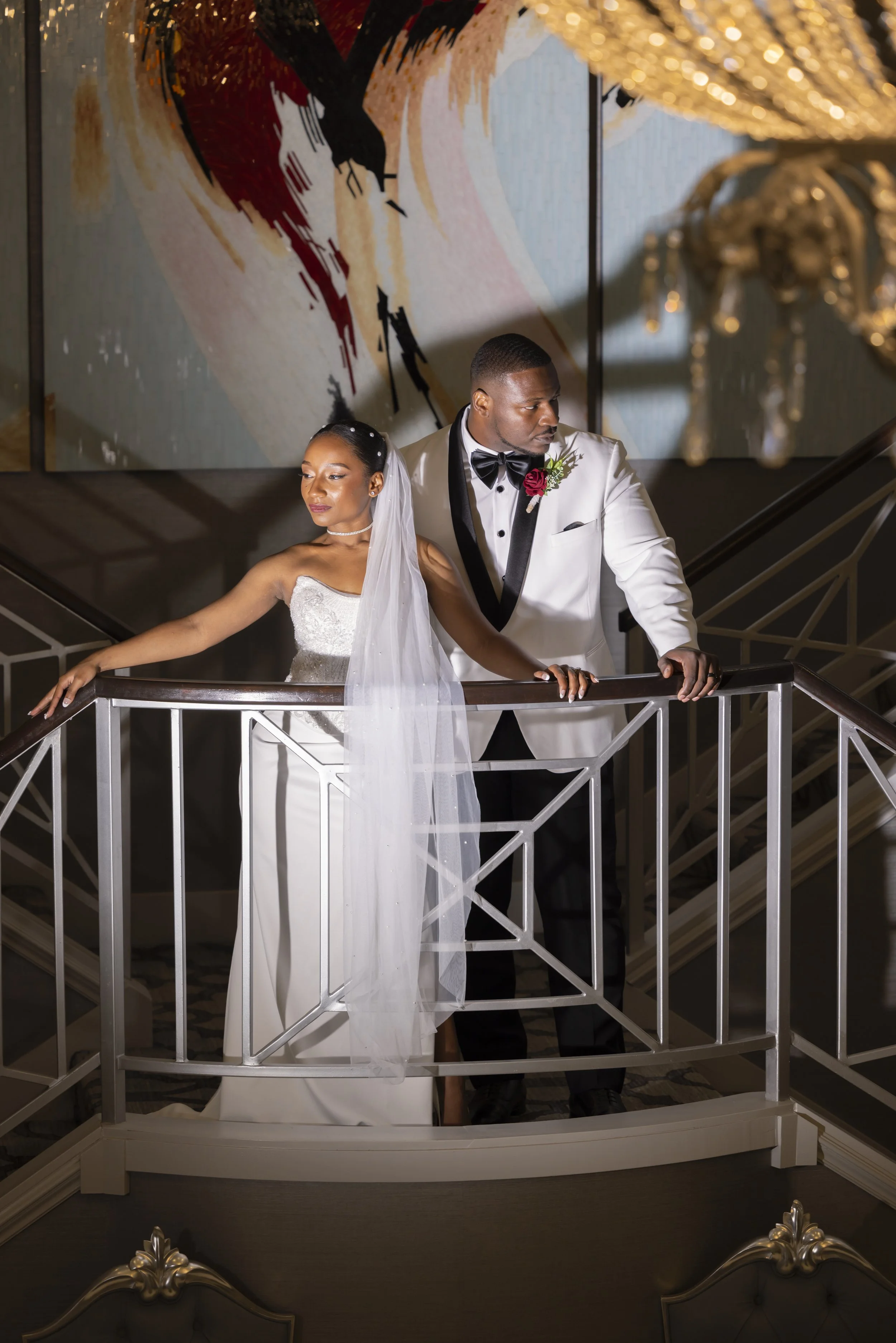 A bride in a white wedding gown and veil and a groom in a white tuxedo with a black bow tie pose on a staircase at a wedding venue, with a large abstract painting in the background and a chandelier overhead.