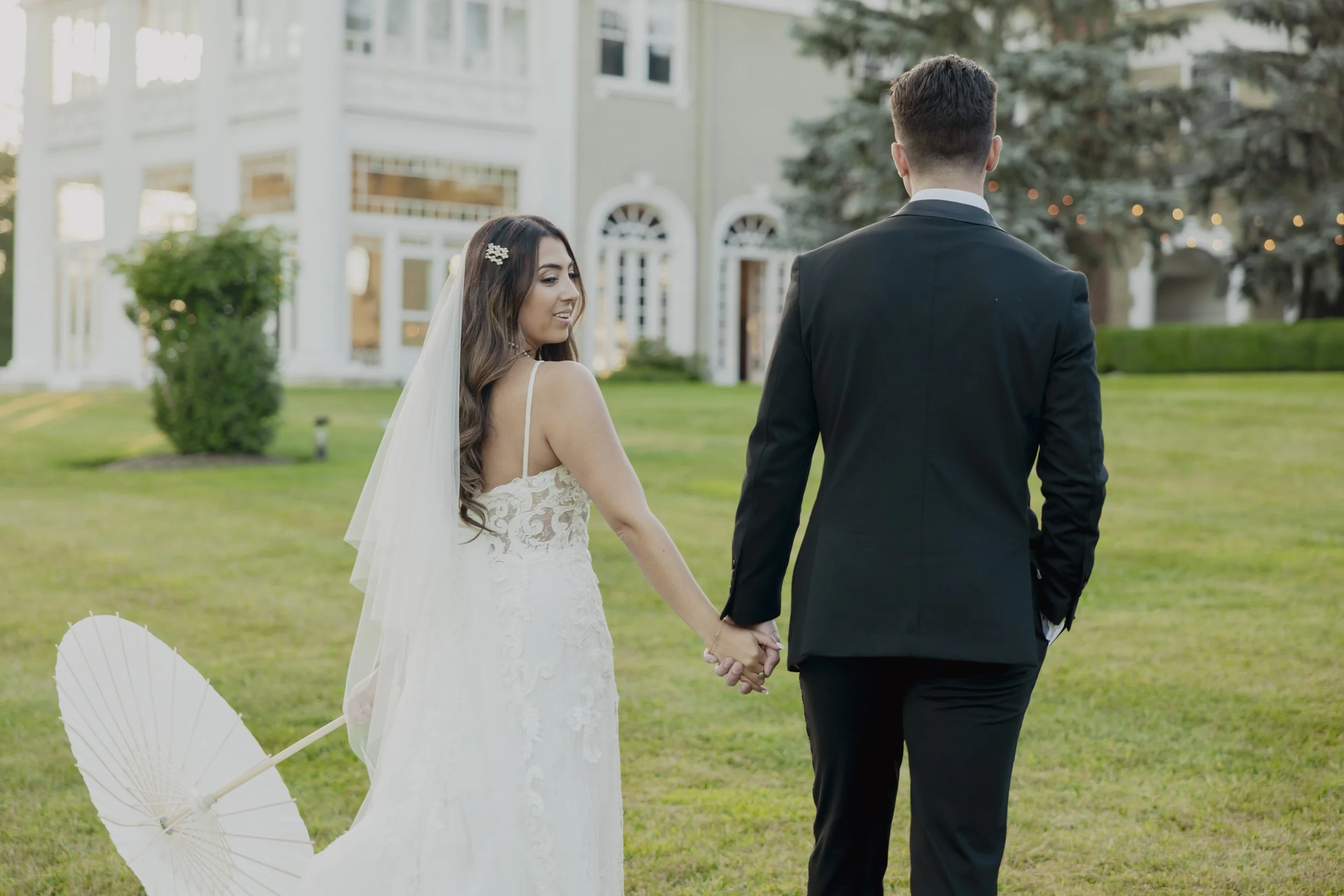 A bride and groom holding hands and walking on a lawn in front of a large house during their wedding. The bride is wearing a white lace wedding dress and veil, and the groom is dressed in a black suit.