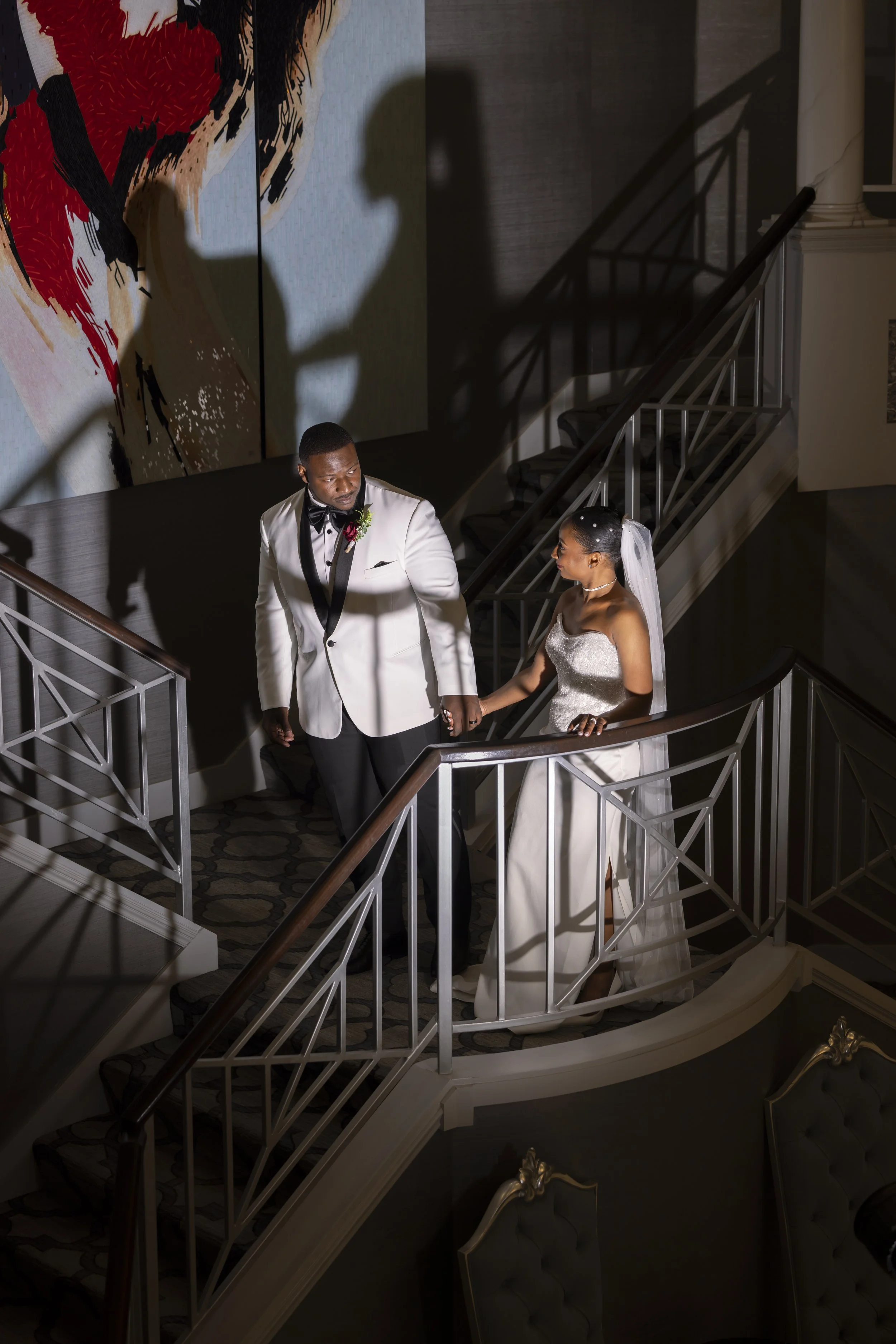 A bride and groom standing on a staircase holding hands, with a shadow of a kiss projected on the wall behind them, during their wedding celebration.