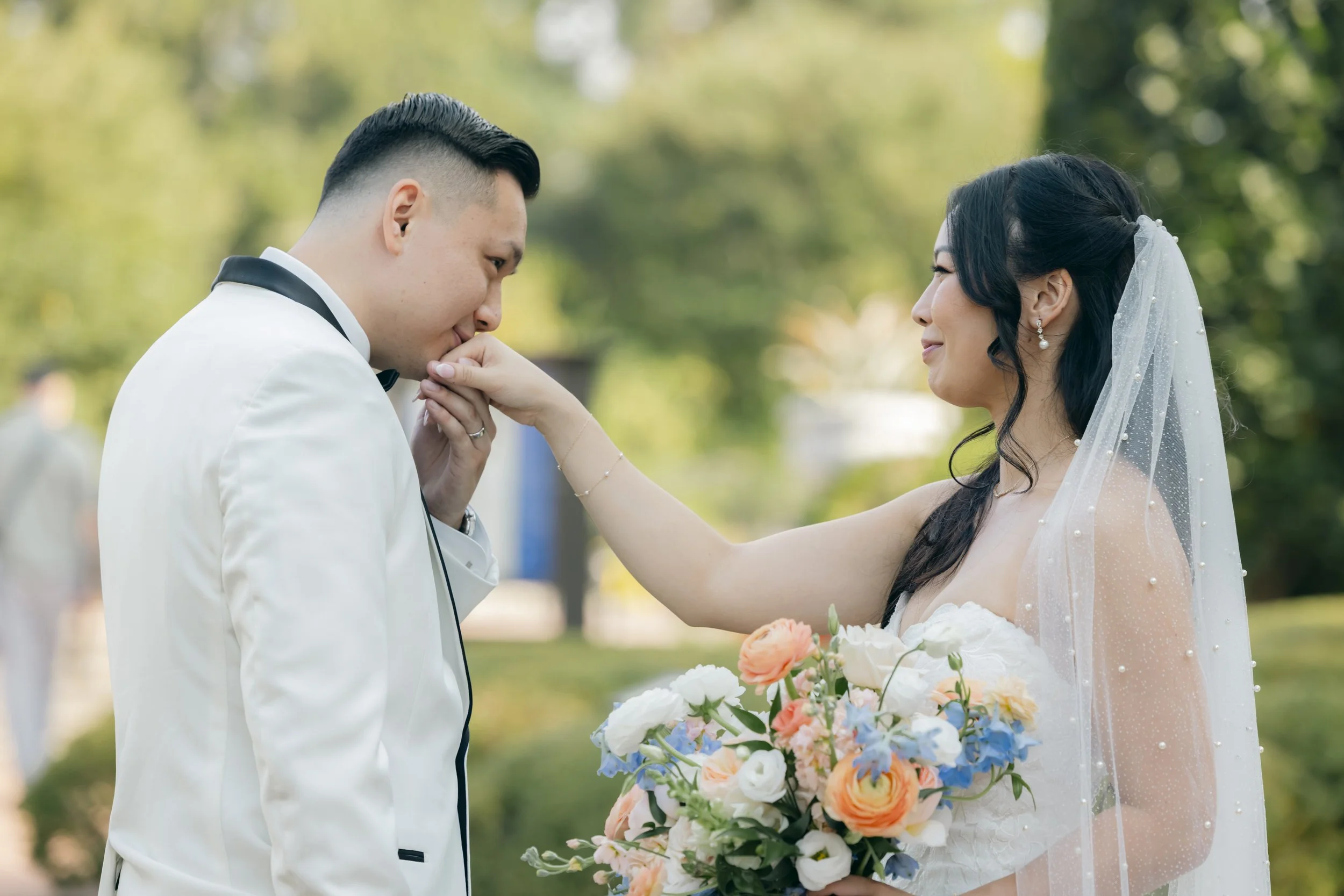 A bride in a wedding dress holding a bouquet of flowers is touching a groom's lips, who is in a white tuxedo, during an outdoor wedding ceremony.