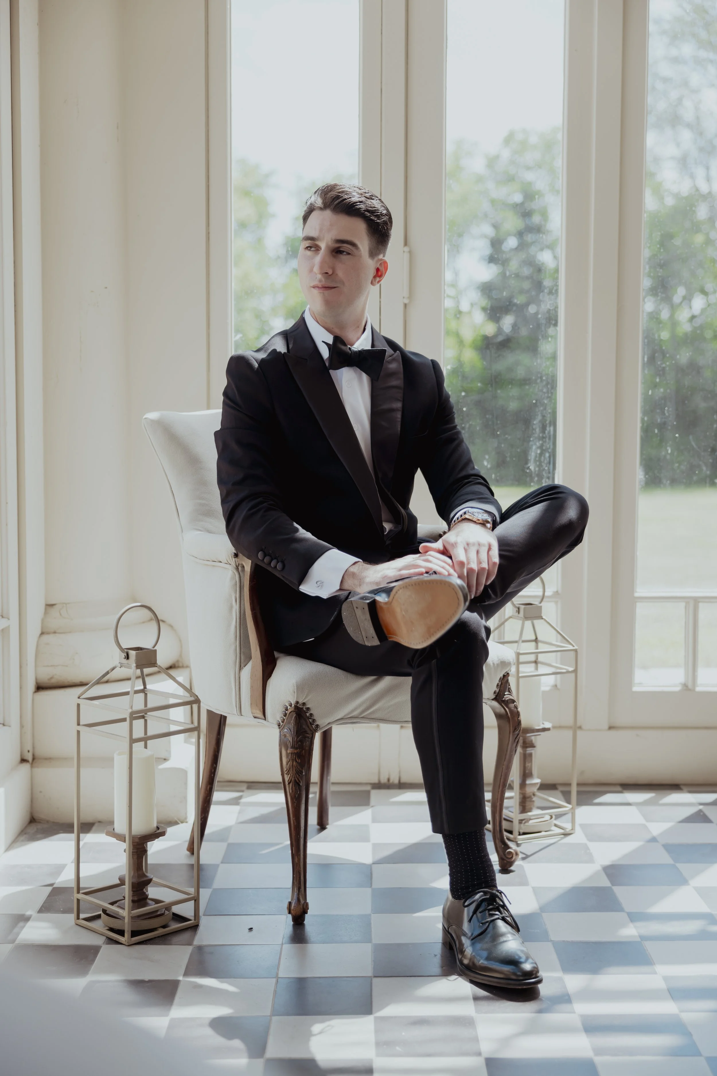 A young man in a tuxedo sitting on a vintage armchair, adjusting his shoe, in a bright room with large windows and checkered floor.