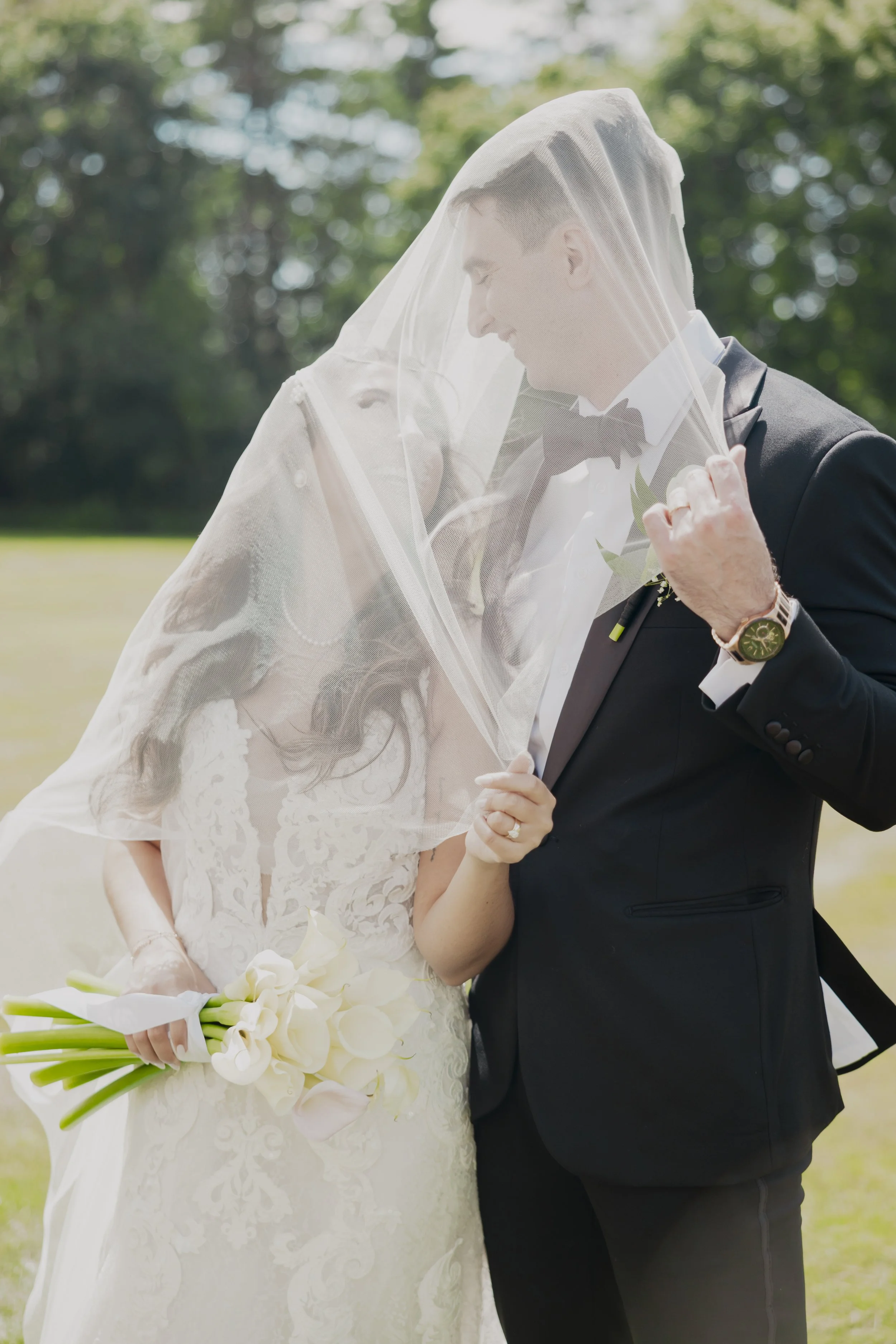 A bride and groom are standing outdoors, sharing a romantic moment under a veil. The bride is holding a bouquet of white flowers, and both are smiling with their eyes closed.