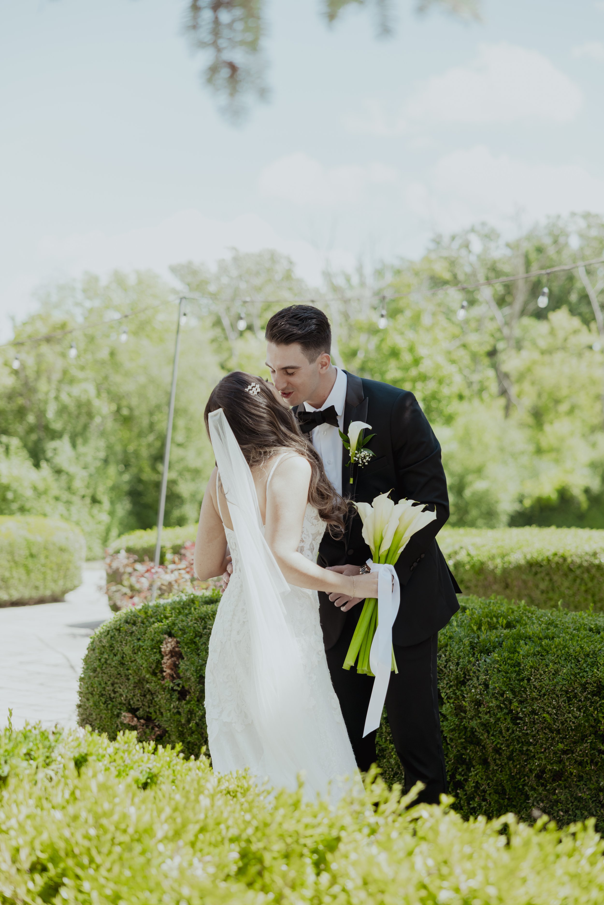 A bride and groom share a romantic moment outdoors among green bushes and trees, with string lights overhead, during their wedding.