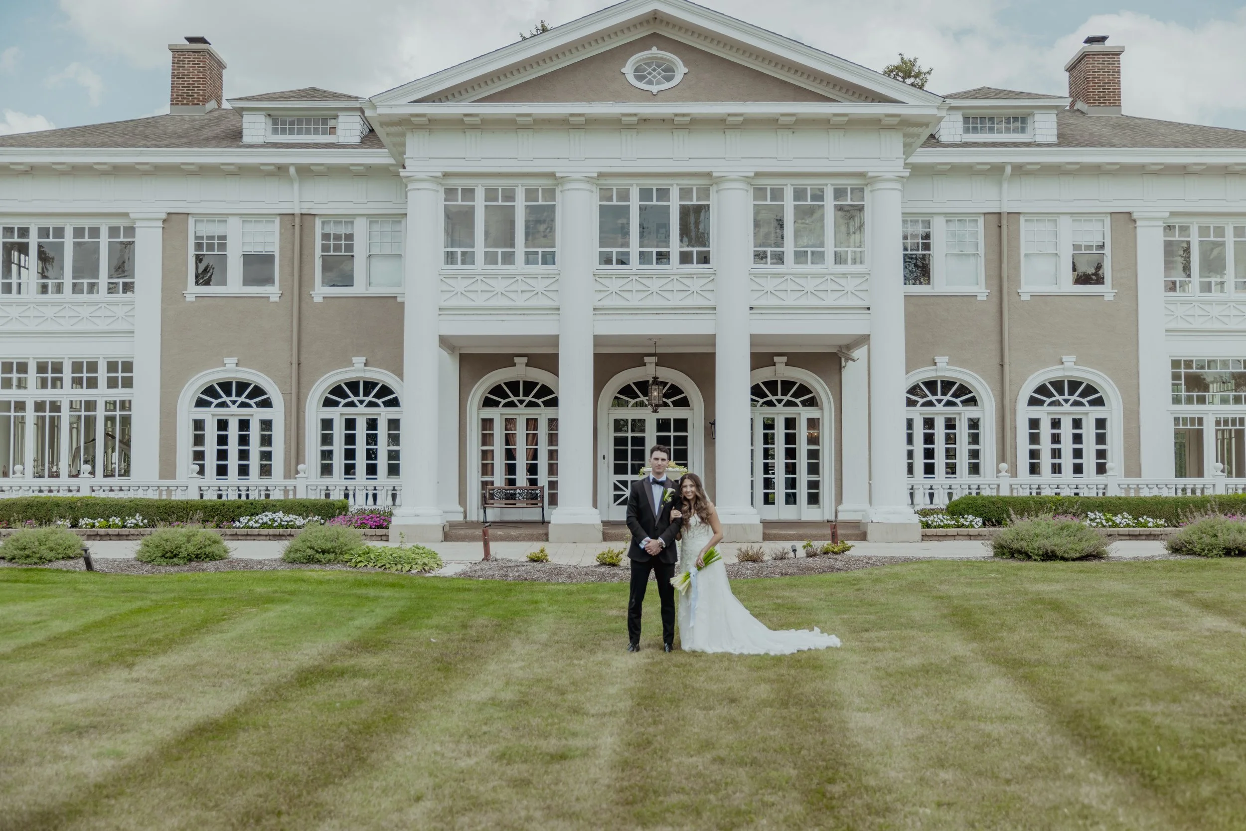 A bride and groom standing on a green lawn in front of a large, elegant house with white columns and multiple large windows, during daytime.