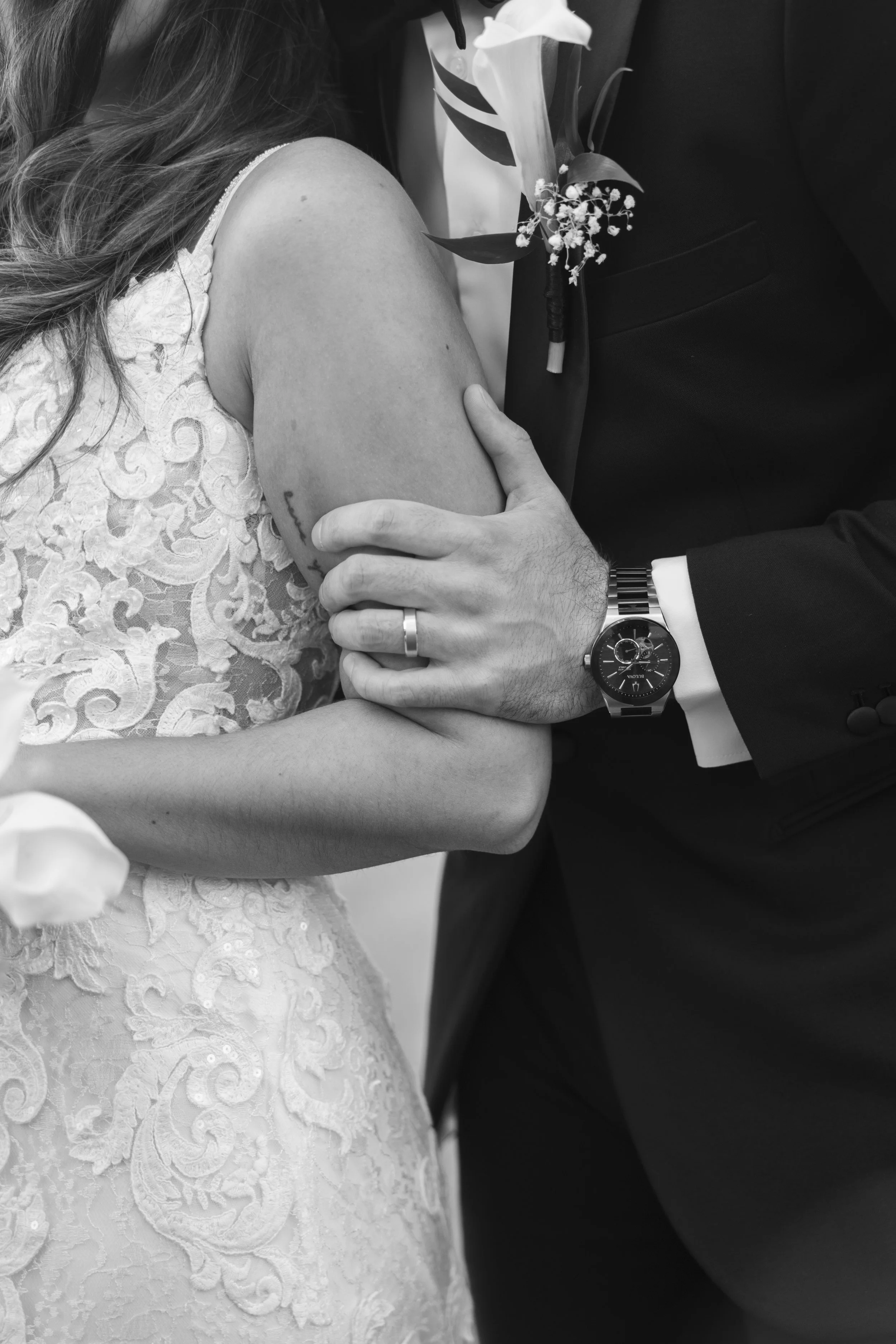 A black and white photo of a bride and groom embracing. The bride wears a lace wedding dress, and the groom in a tuxedo with a boutonniere. The groom's arm with a watch and wedding band is around the bride.