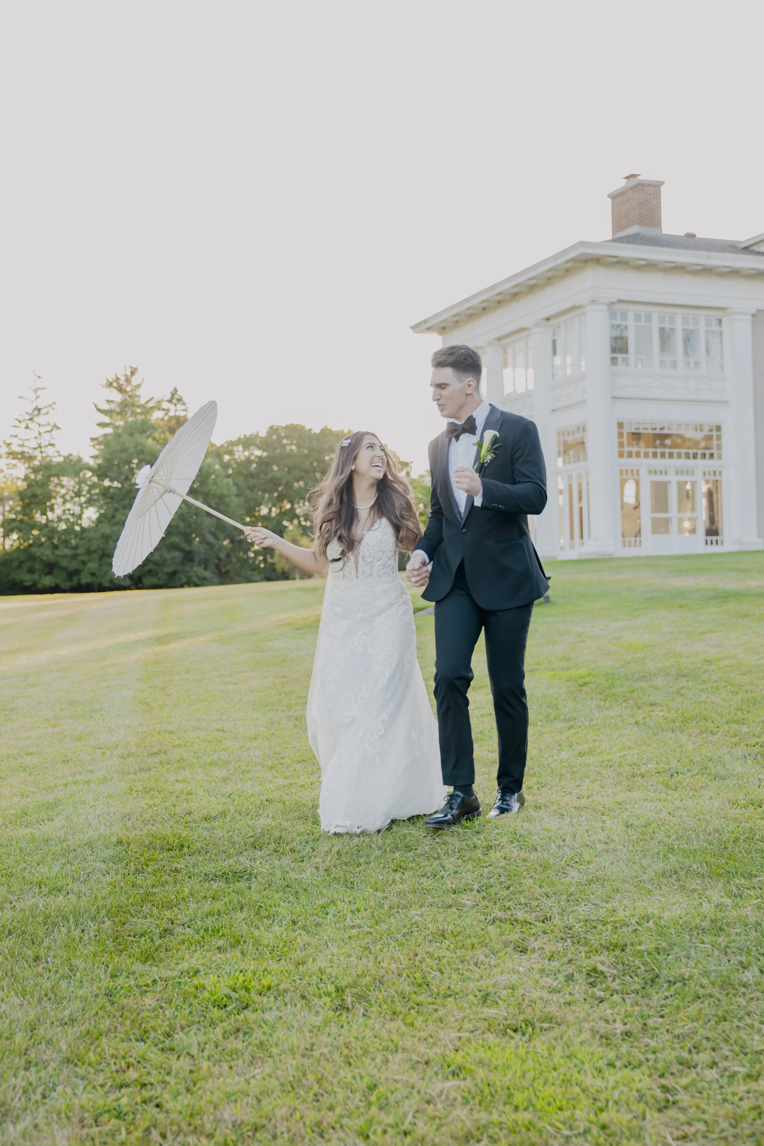 Bride and groom walking on grass lawn beside a large white house, holding an umbrella, smiling.