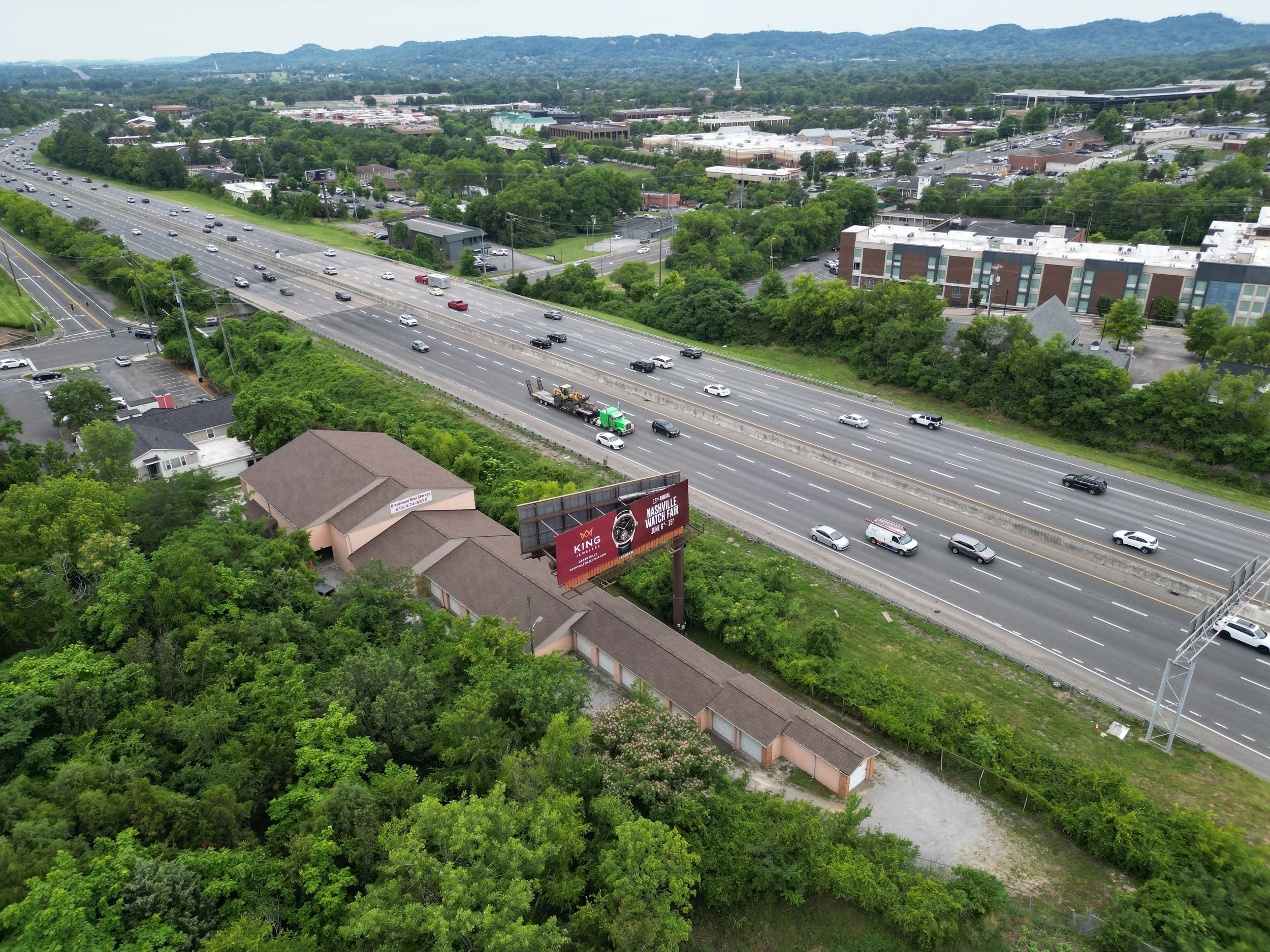 Aerial view of a highway with multiple lanes and cars driving in both directions, surrounded by greenery and trees. Nearby, there is a large billboard and several buildings. In the background, there are more buildings, roads, and distant hills.