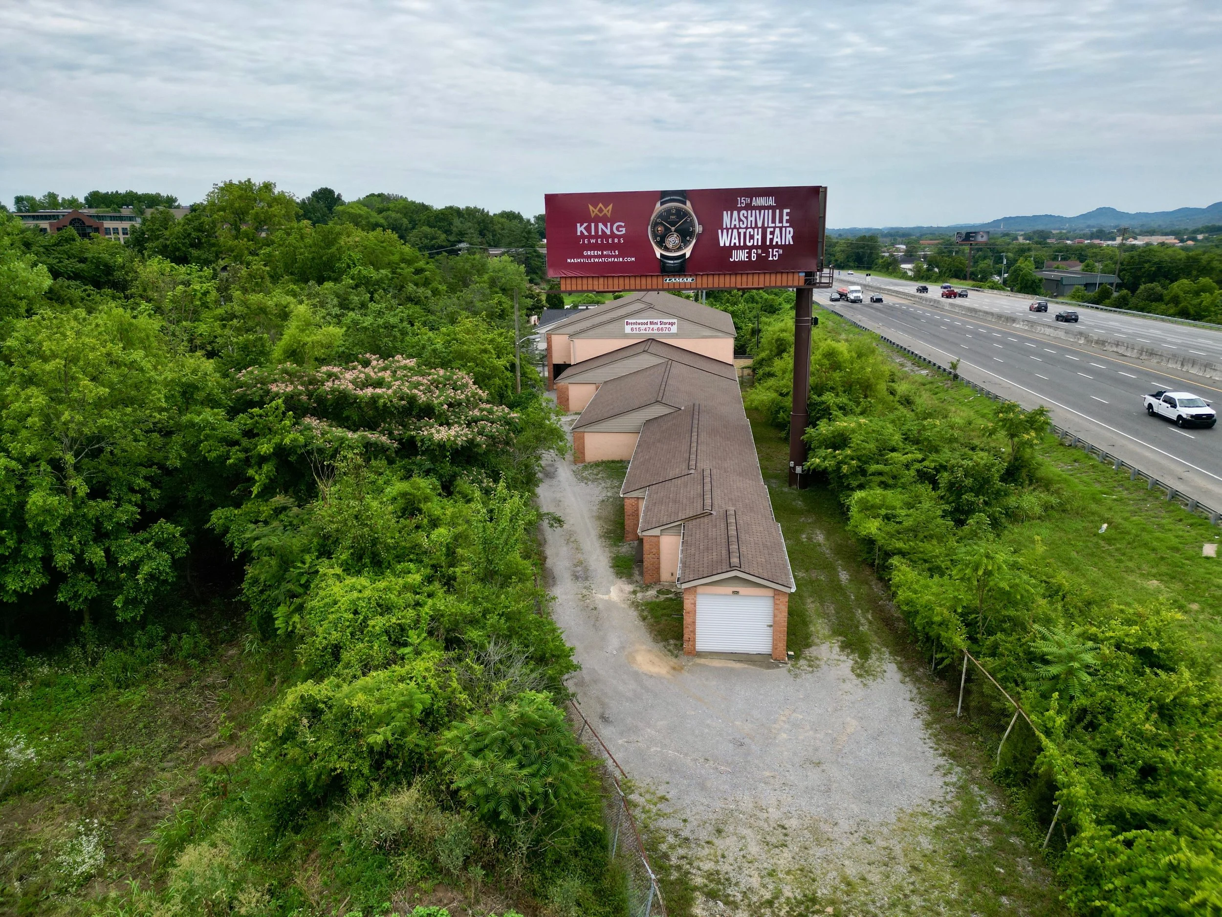 Aerial view of a storage facility with several units, surrounded by green trees. A large billboard advertising the Nashville Watch Fair is visible near the highway with cars. Overcast sky in the background.
