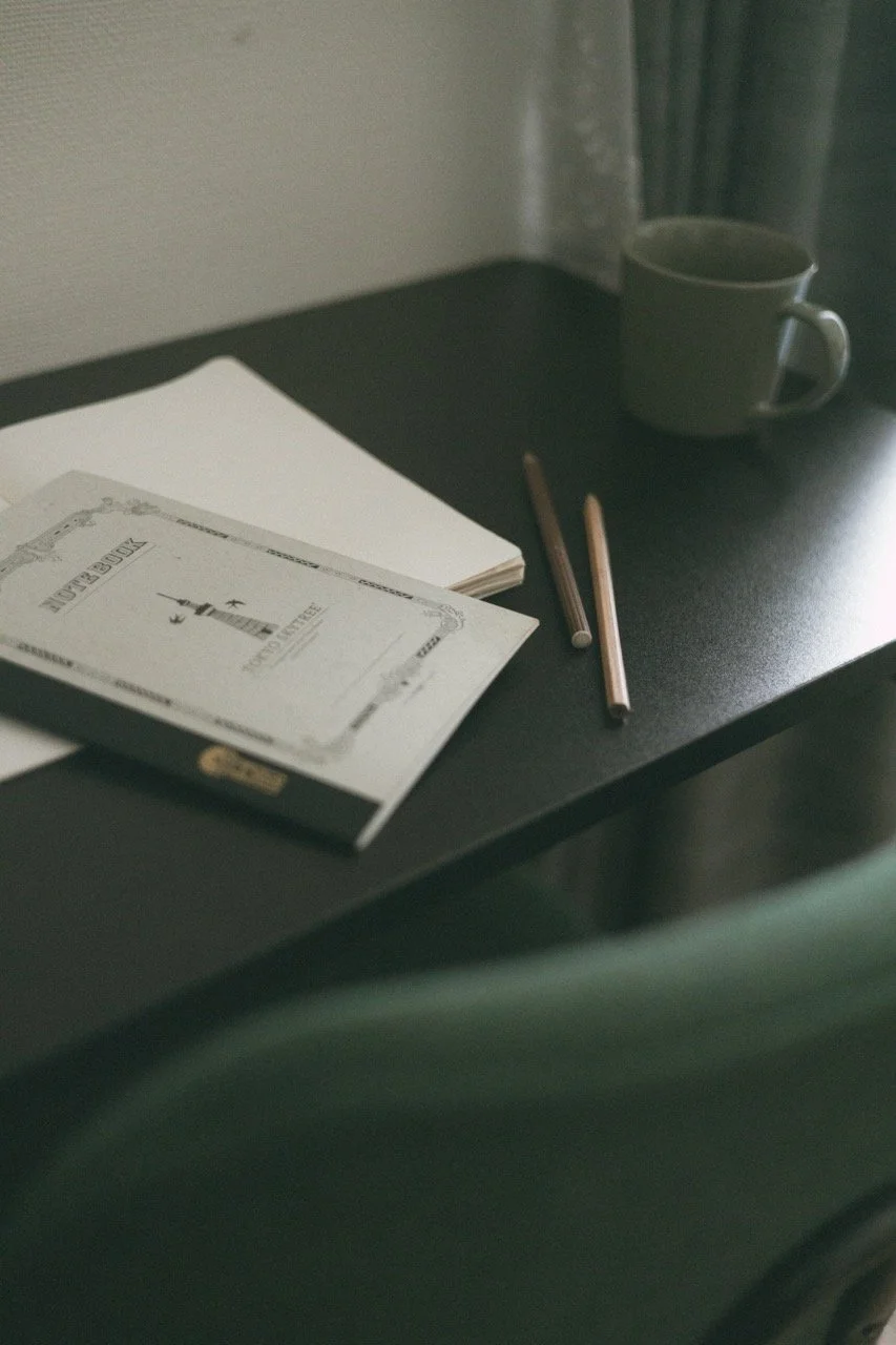 A black desk with a closed book titled "Book Review" featuring a lighthouse drawing, an open notebook, two pencils, and a green mug with a handle.