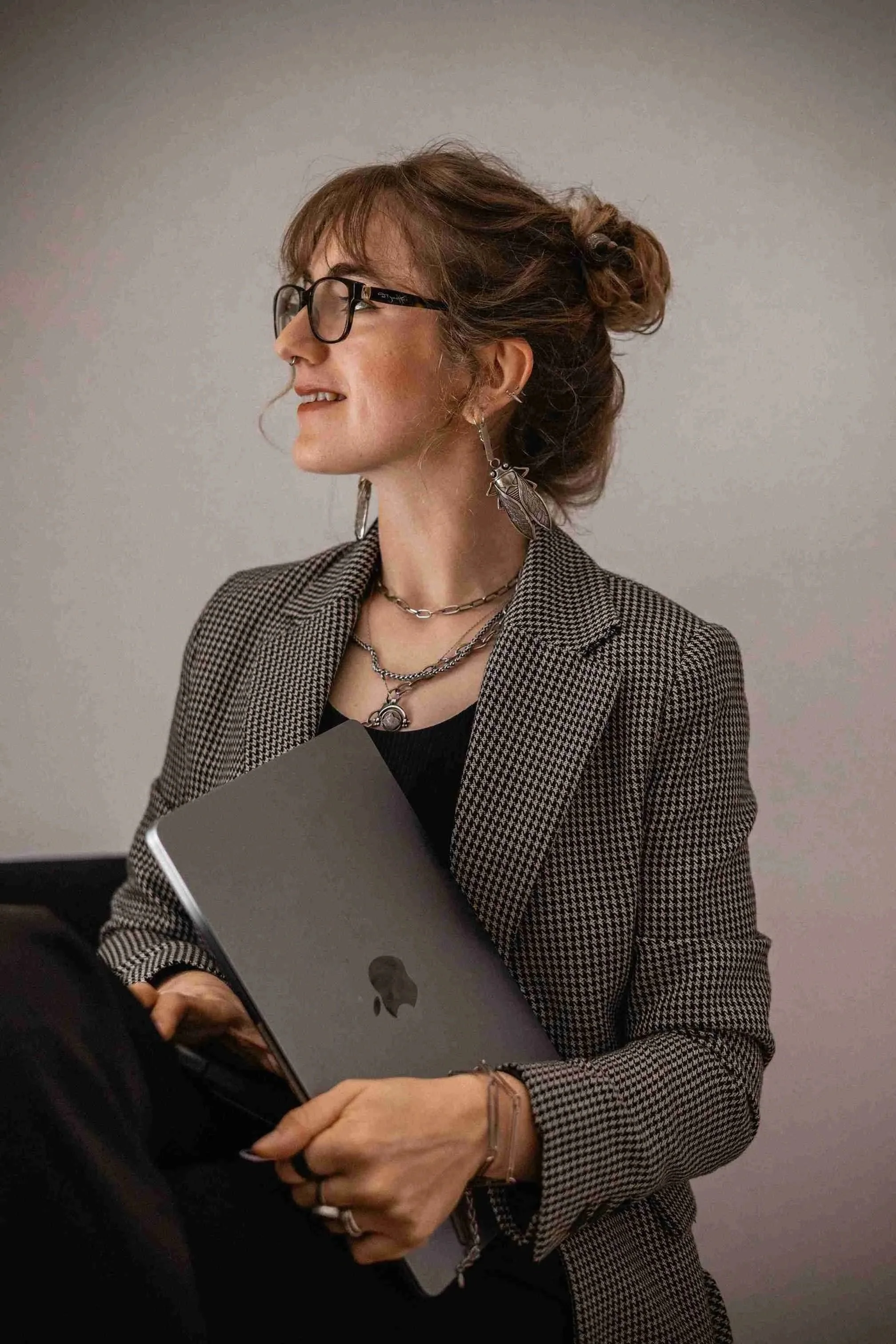 A woman with light brown hair in a bun wearing glasses, silver jewelry, and a houndstooth blazer holding a closed MacBook laptop, standing against a plain background.