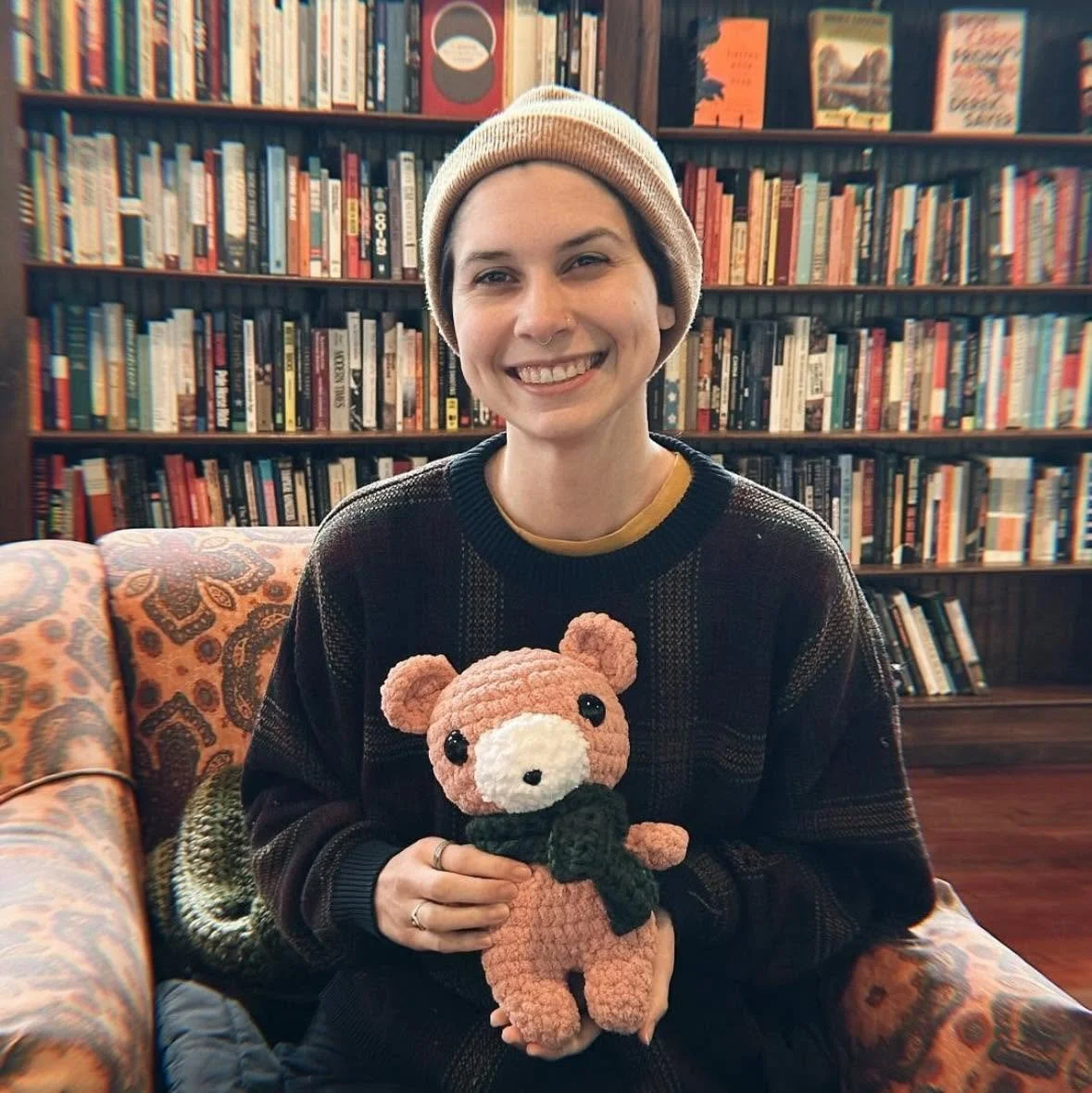 A young woman with short hair, wearing a beanie and a dark sweater, smiling while holding a crocheted teddy bear in a cozy room with a bookshelf filled with books in the background.