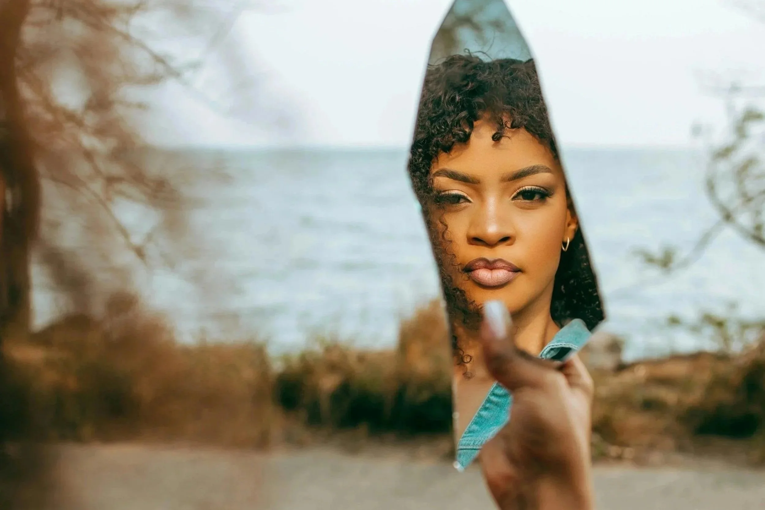 Black woman holding a broken mirror shard reflecting her face, standing near the ocean symbolizing self-reflection, inner healing, and the emotional fragmentation caused by minimized trauma.