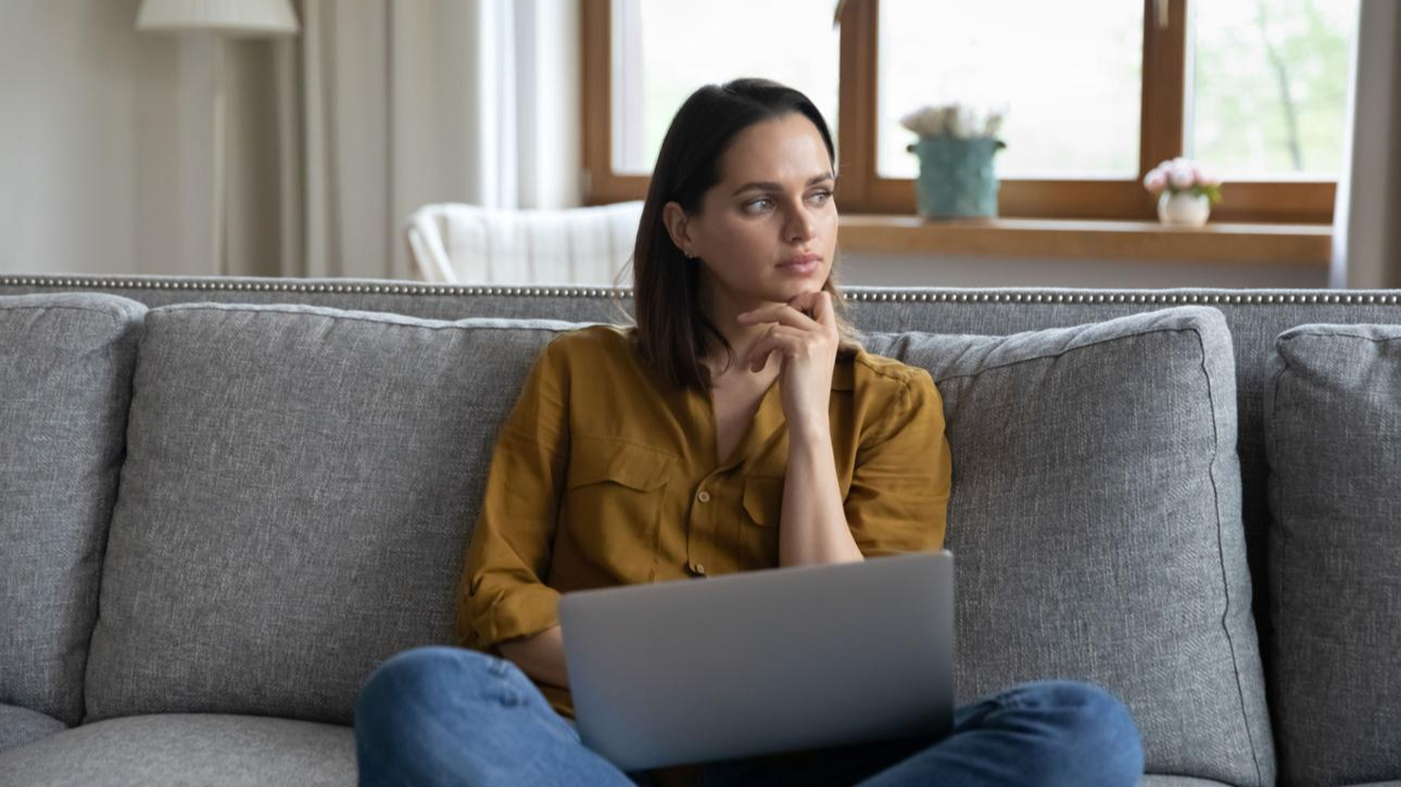 Woman sitting on a couch with a laptop during a quiet moment of reflection at home.