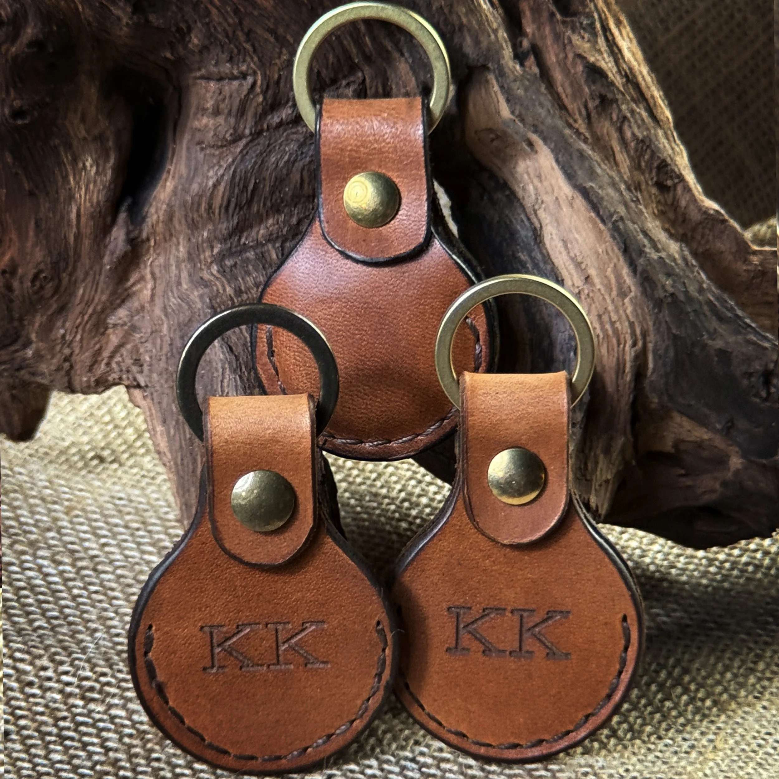 Three leather keychains with brass rings and the initials "KK" embossed, hanging from a wooden branch.