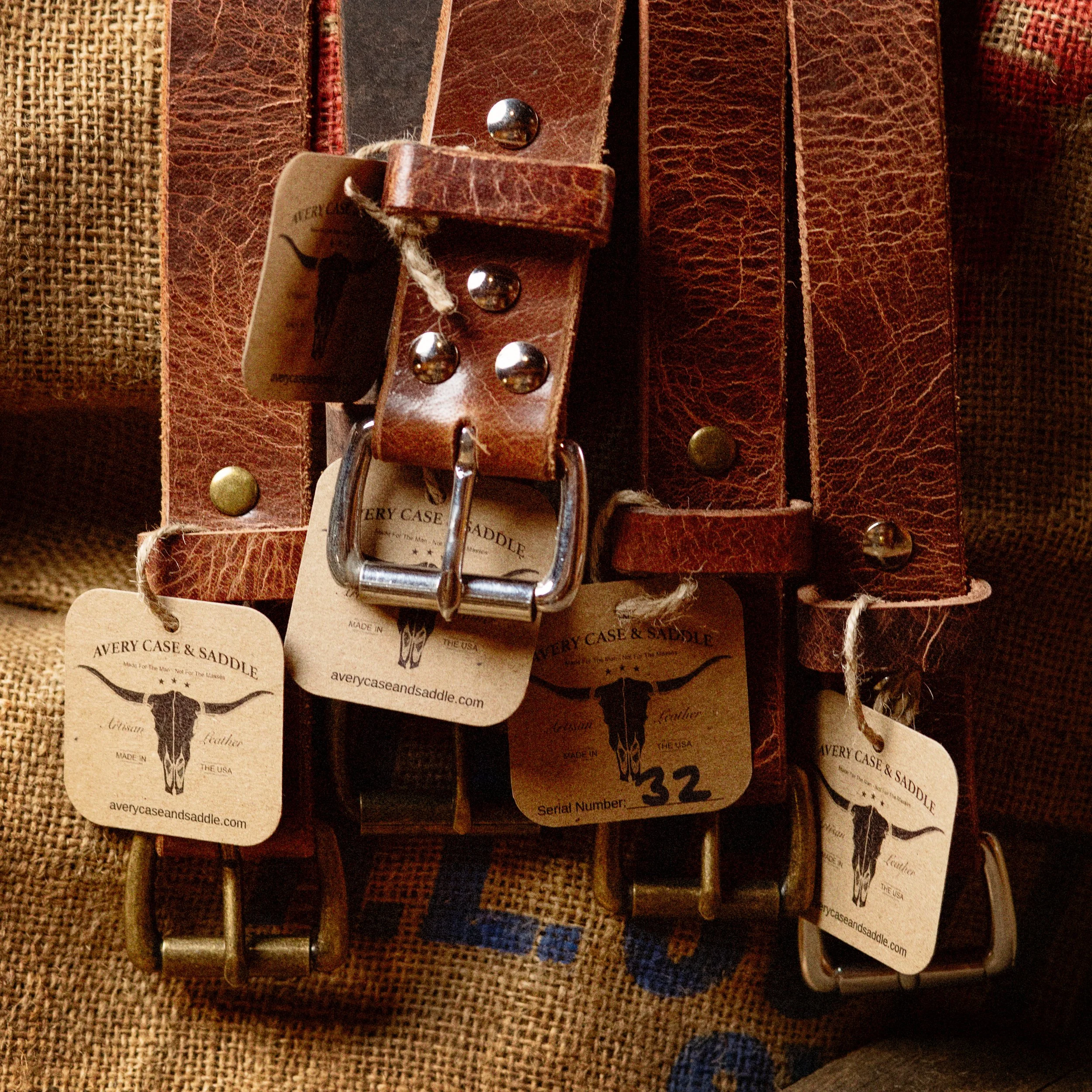 Close-up of three brown leather belts with metal buckles hanging on burlap fabric, each labeled with tags from Avery Case & Saddle.