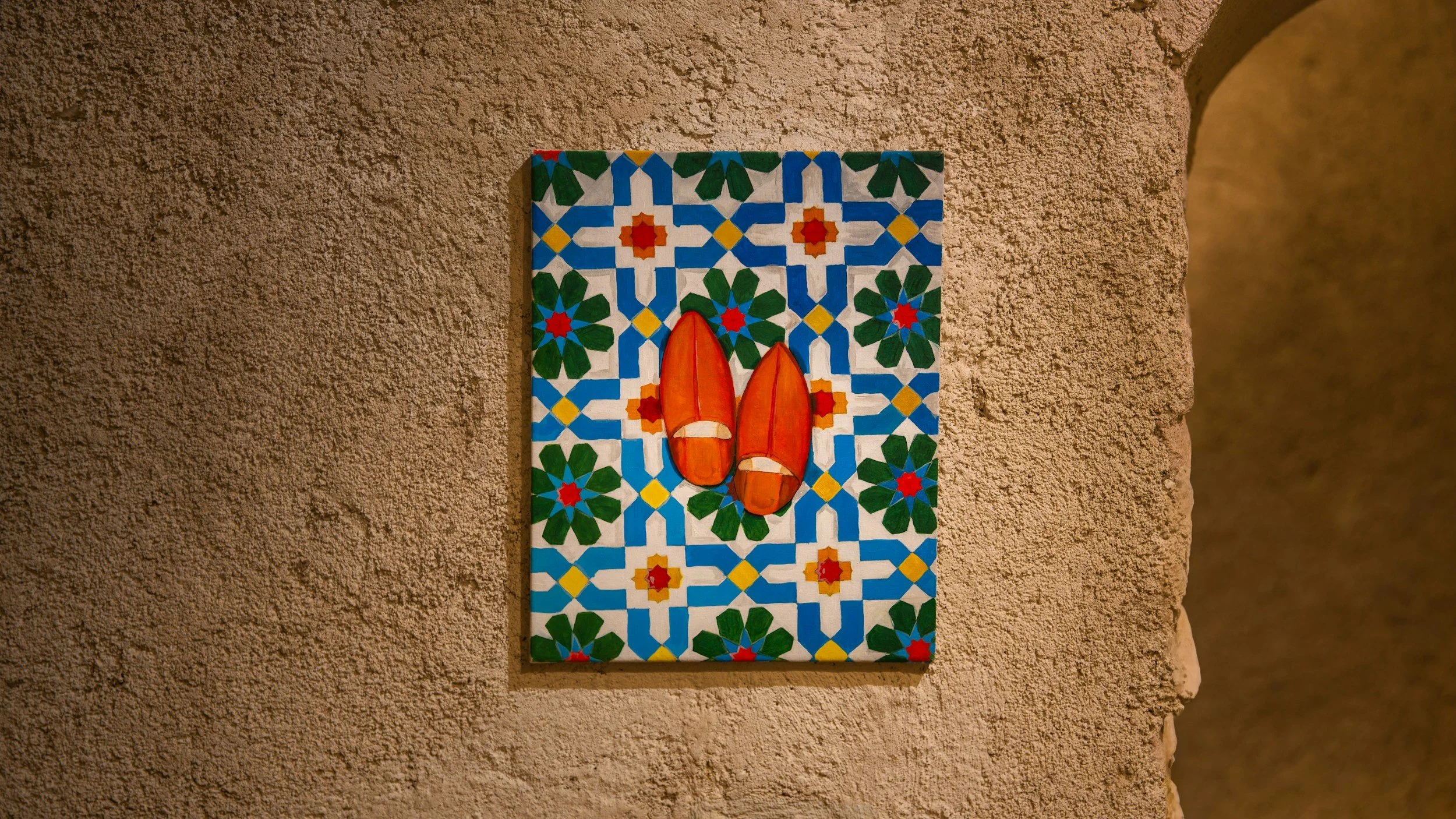 Painting of red shoes on colorful geometric tile background mounted on a textured beige wall.