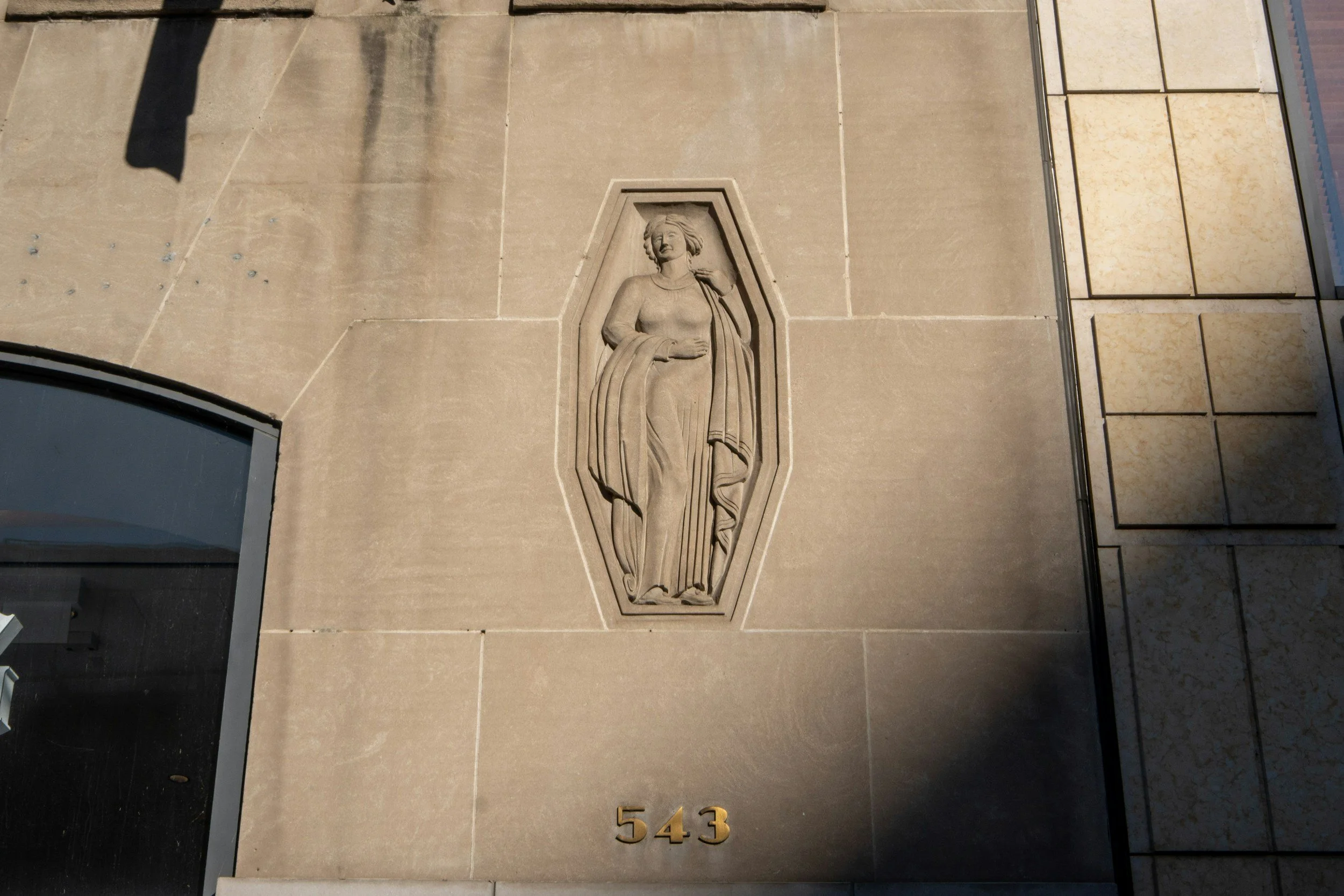 Stone relief of a woman in a flowing dress with a draped shawl, located above the number 543 on a beige building wall. Unlocking the hidden wisdom behind numerology and centuries of spirtiual realms.