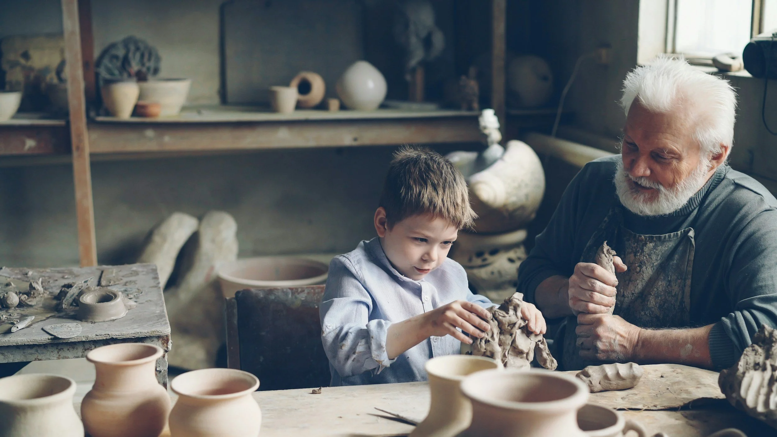 A young boy and an older man, possibly a potter, working together on clay in a pottery studio. Various clay pots are on the table in front of them, and shelves with pottery and tools are in the background. Appreciating the journely of discovery.